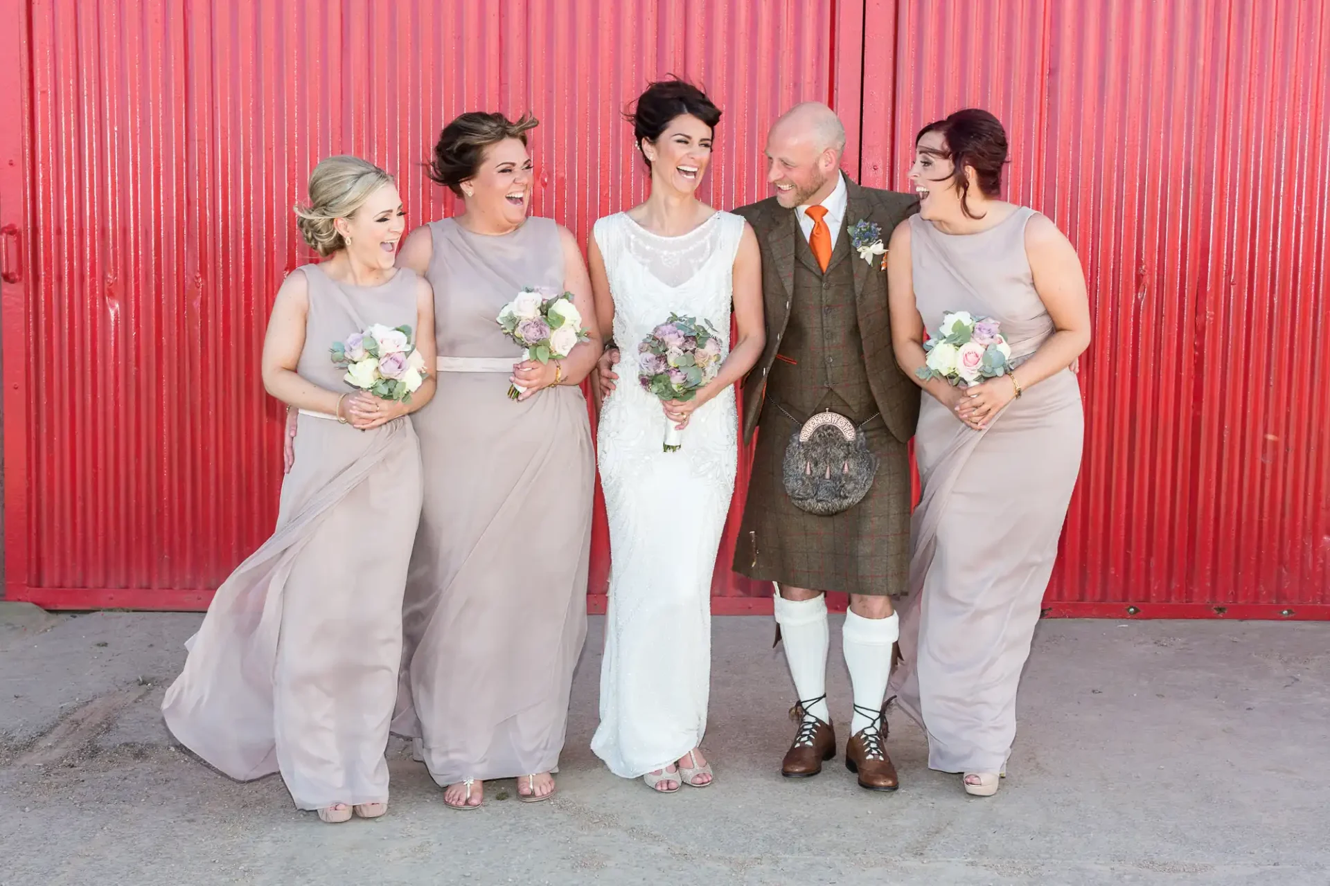 Bride and groom in traditional scottish attire laugh with three bridesmaids against a red background.