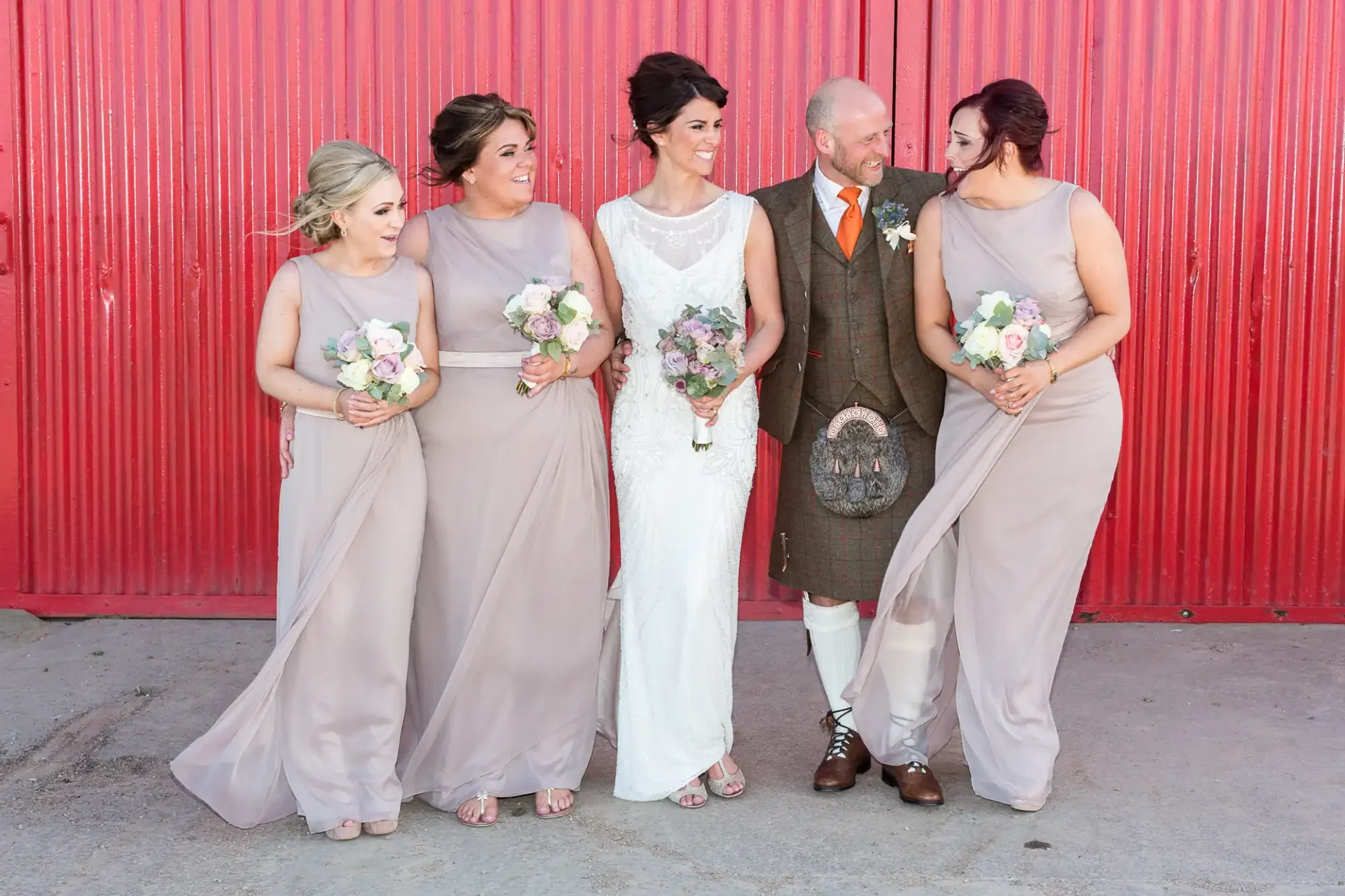A bride in white and three bridesmaids in taupe dresses laugh with a bearded groom in a traditional kilt, standing in front of a red wall.