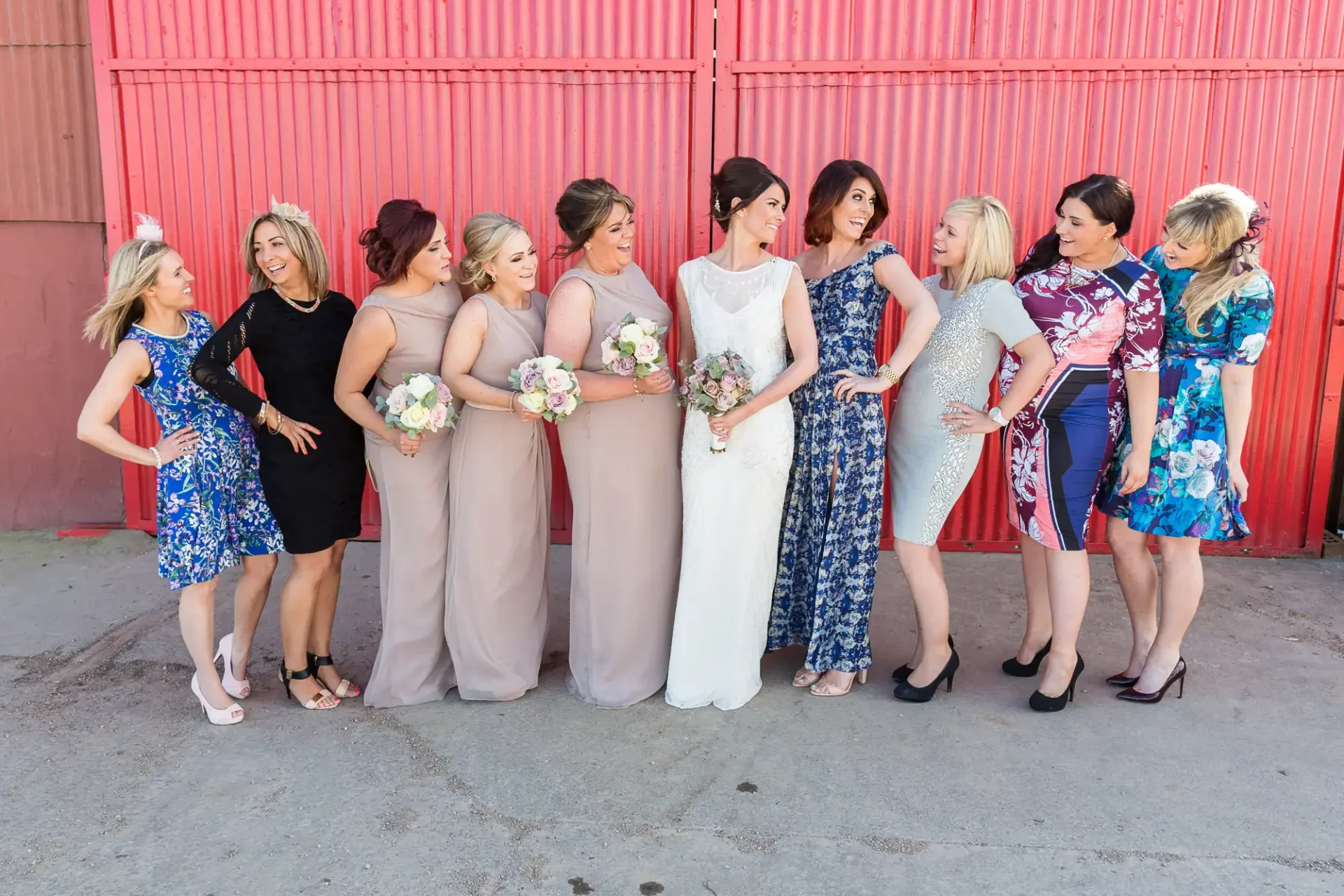 A group of women in elegant dresses posing with a bride against a red corrugated metal background.