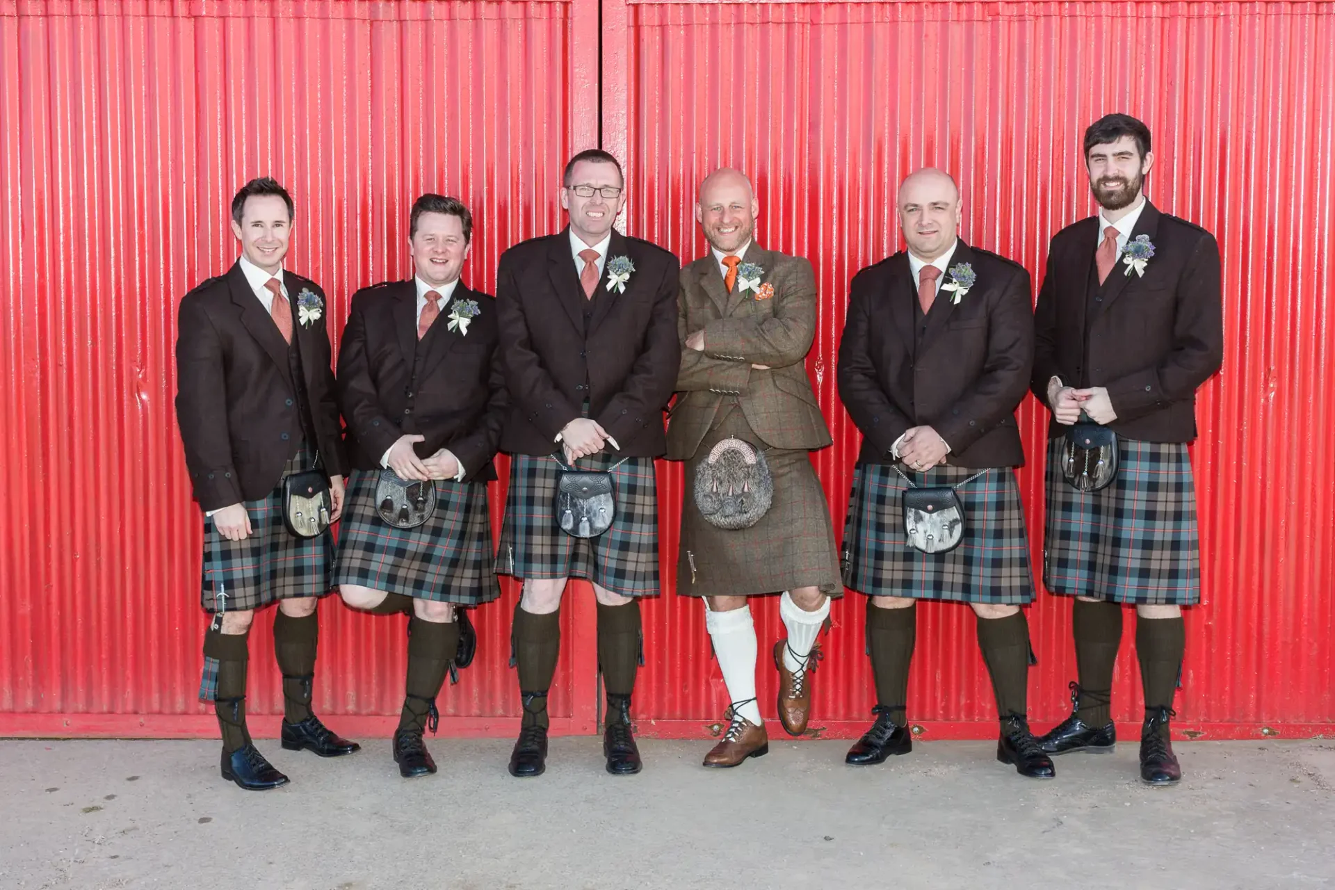 Six men in traditional scottish kilts and jackets standing in front of a red corrugated metal wall, smiling for the camera.