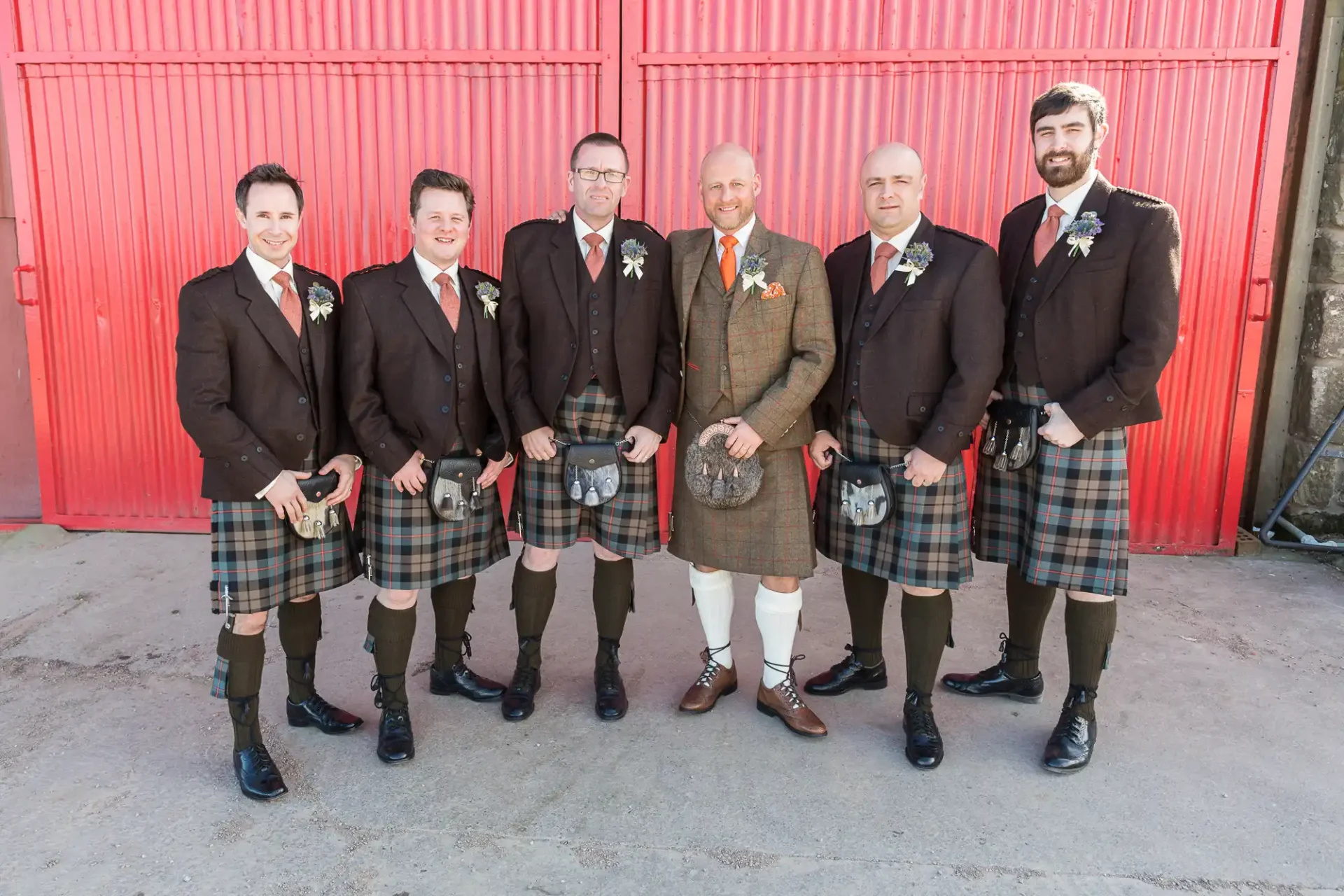 Six men wearing traditional scottish kilts and jackets pose for a photo in front of a red barn door.