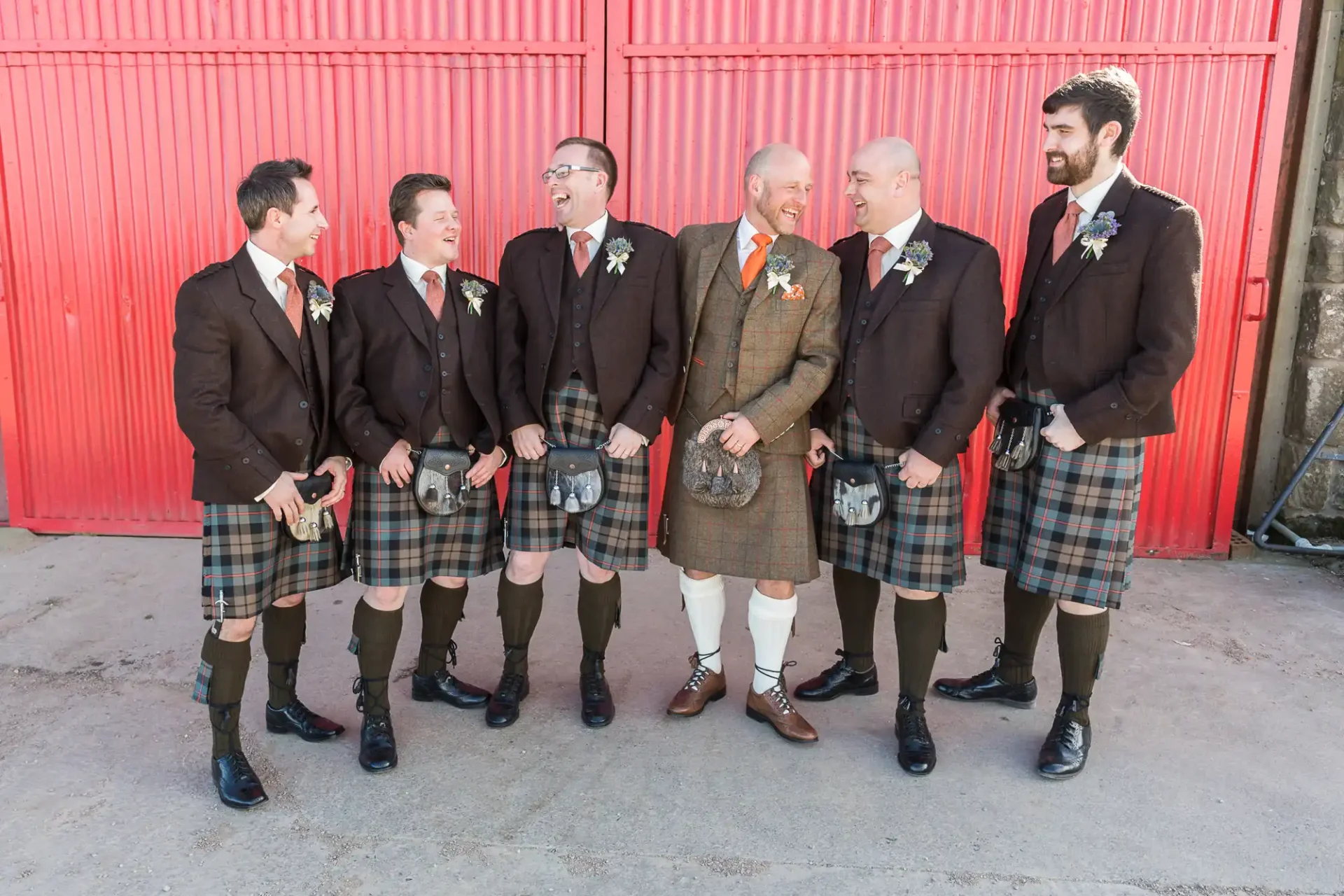 Six men in traditional scottish kilts and jackets, laughing together in front of a red barn door.