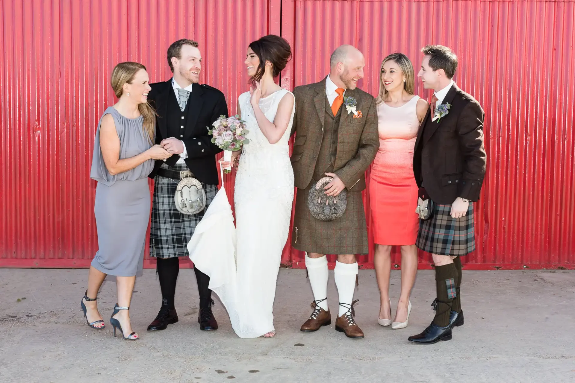 Group of six people in formal attire, featuring kilts and a wedding dress, smiling and interacting in front of a red backdrop.