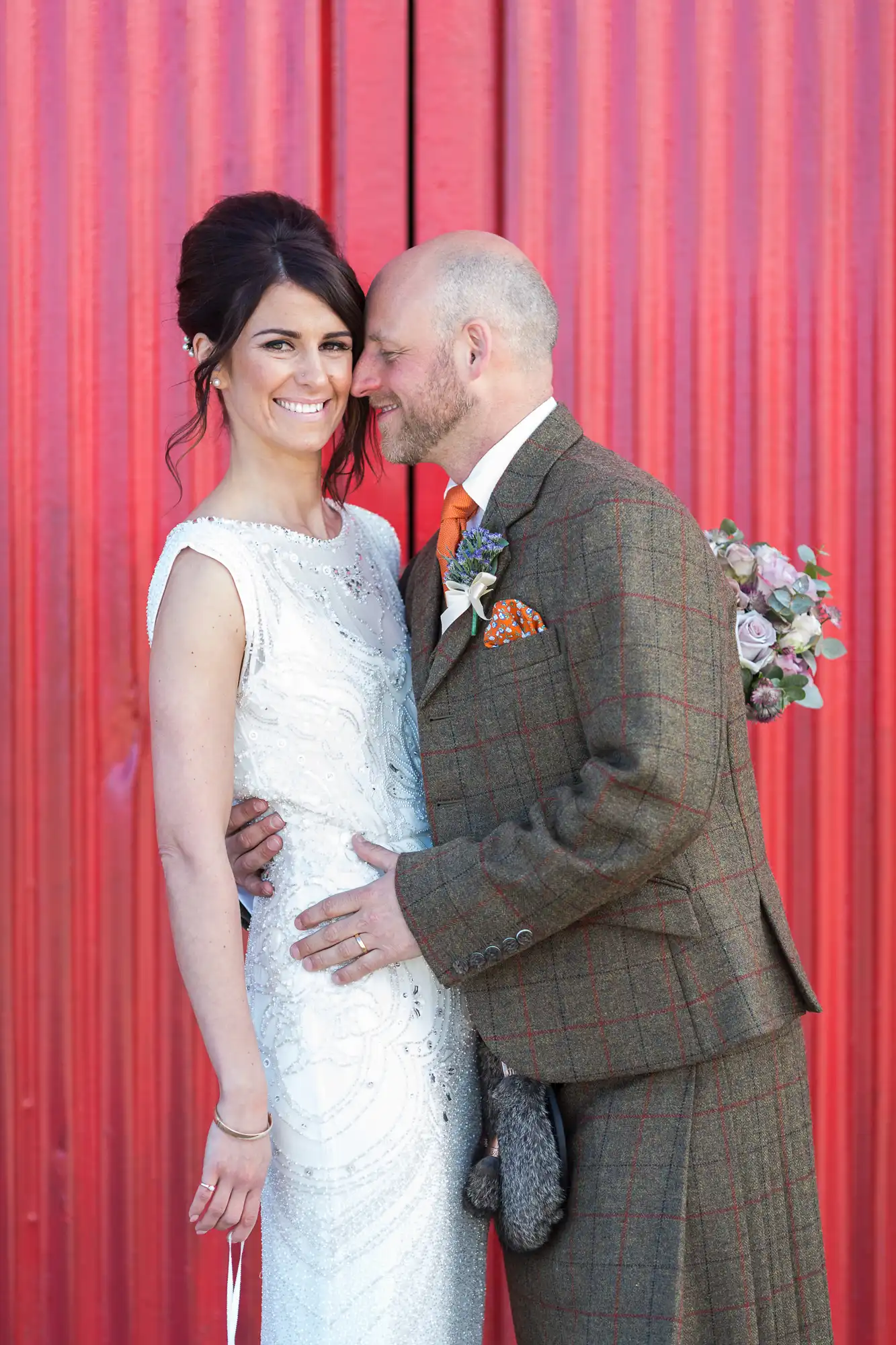 A bride and groom smiling, with the bride in a white dress and the groom in a tweed suit, standing before a red background.