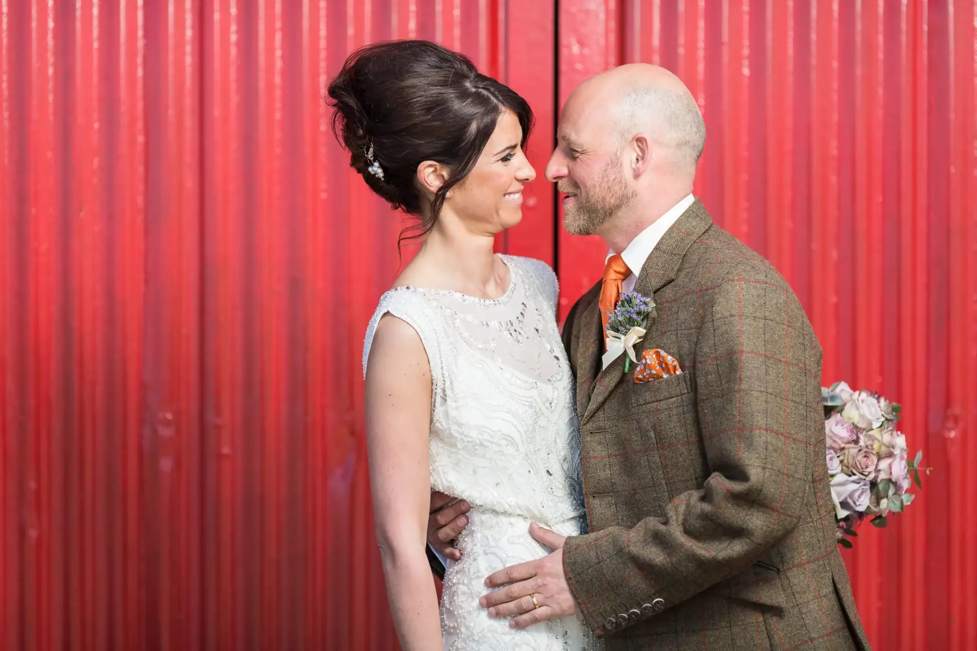A bride and groom smiling at each other, the bride in a white dress and the groom in a tweed suit, against a red corrugated metal background.