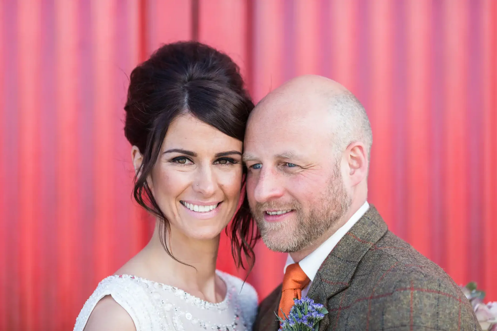A smiling bride and groom posing in front of a red corrugated metal wall, the bride in a white dress and the groom in a tweed suit.