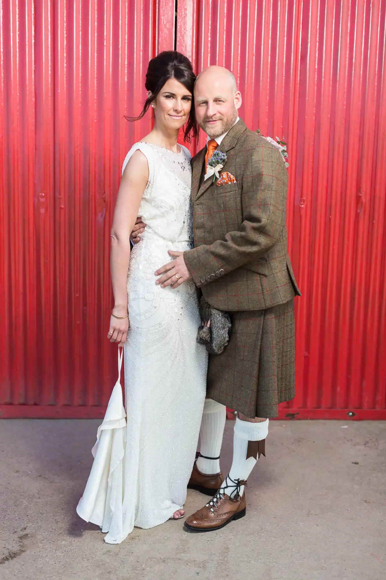 A bride in a white dress and a groom in a traditional scottish kilt and tweed jacket pose in front of a red corrugated metal wall.