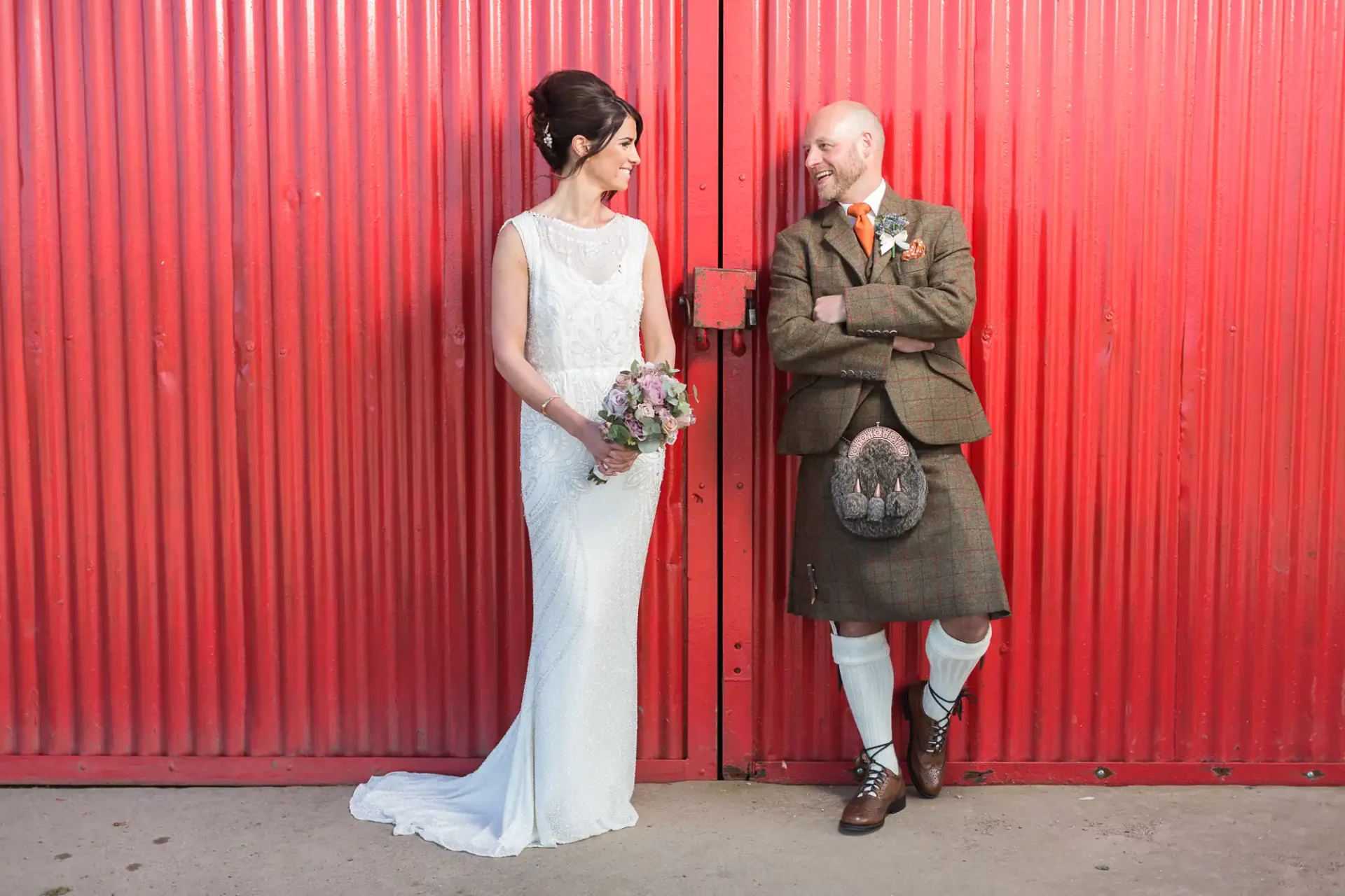 A bride in a white dress and a groom in a kilt sharing a cheerful moment against a vibrant red background.