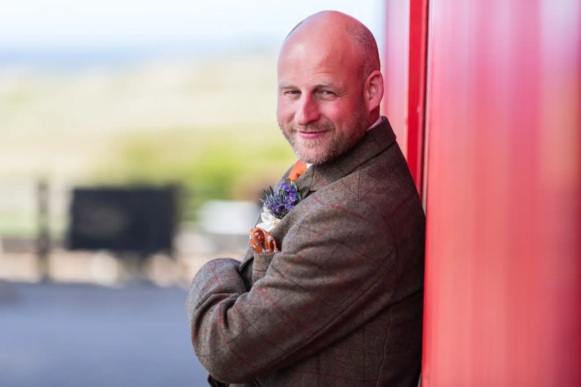 A bald man in a tweed jacket smiling and leaning against a bright red wall, holding flowers.