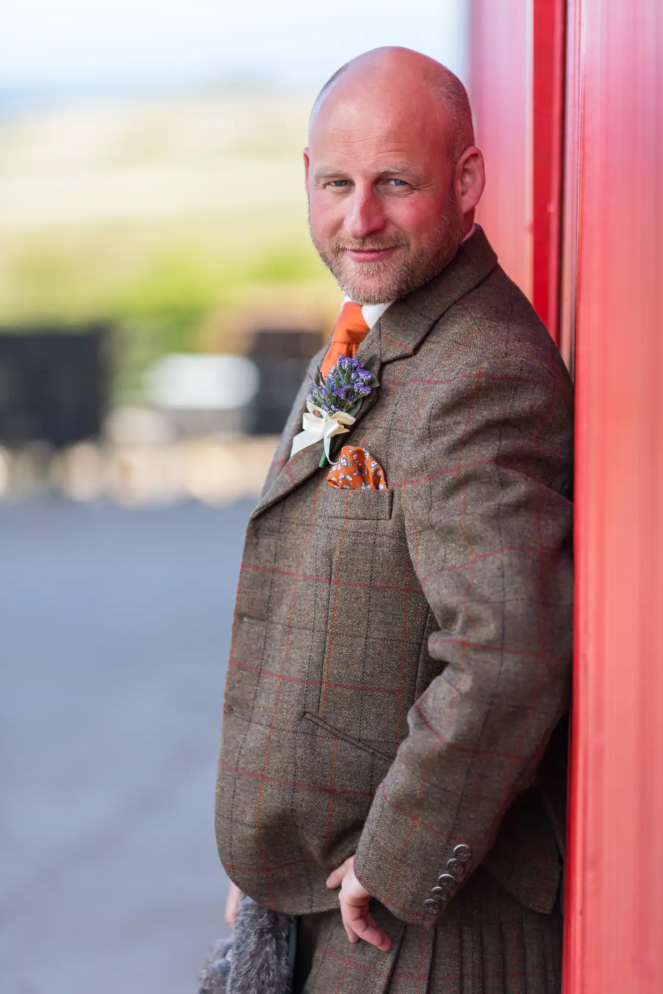 A man in a tweed suit and orange tie leaning against a red door, smiling subtly at the camera.