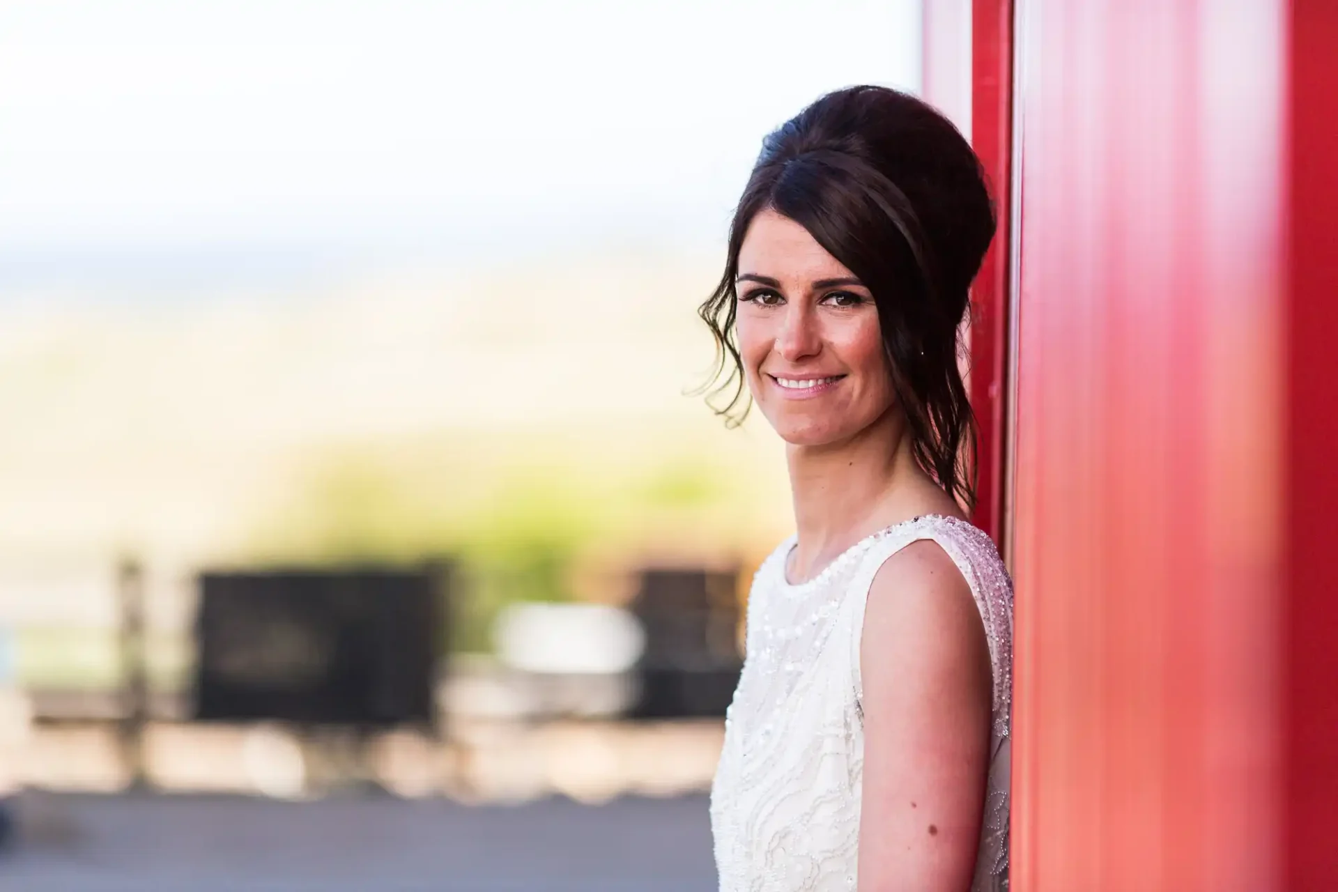 A bride in a white dress smiles gently, standing against a bright red background, with a scenic landscape faintly visible in the distance.