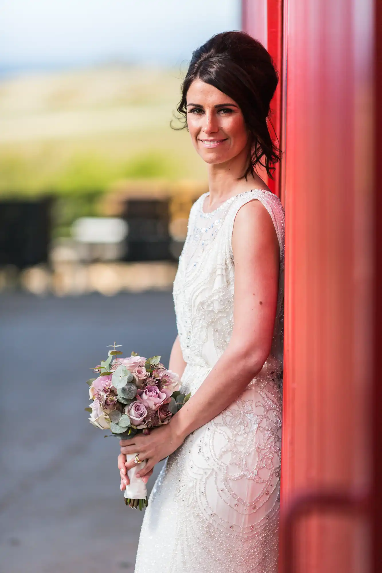 A bride in an elegant white dress, holding a bouquet, smiles subtly next to a vibrant red structure.