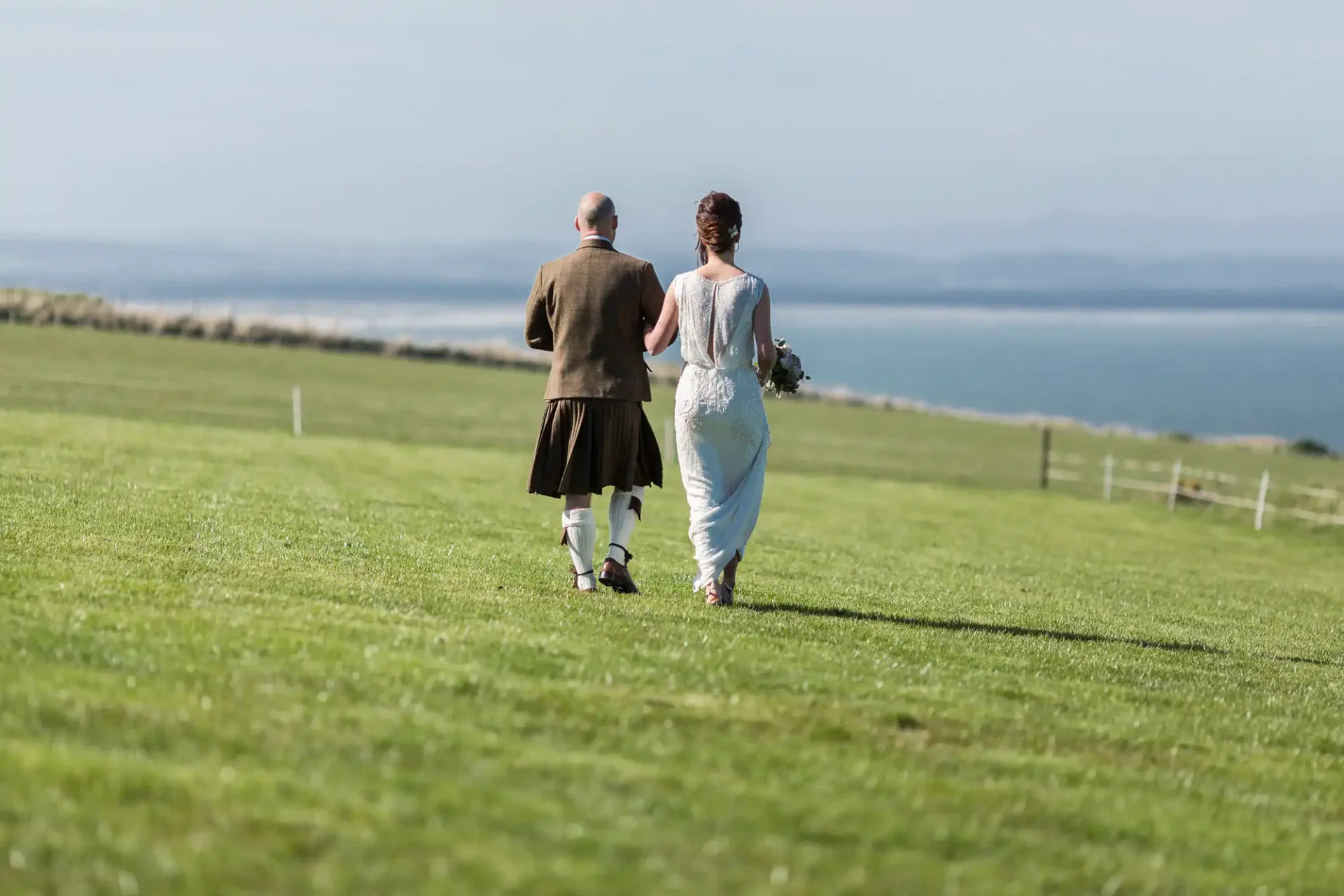 A bride and groom in traditional attire walking on a grassy field by the sea. the groom wears a kilt, and the bride carries a bouquet.