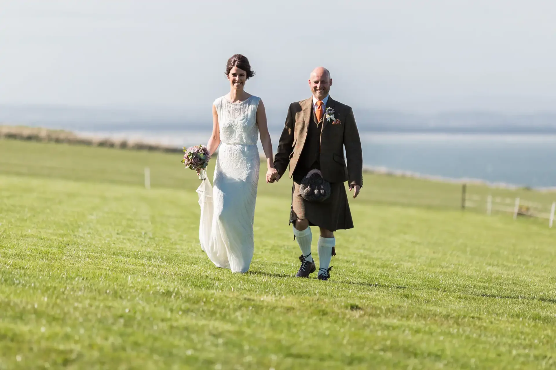 A bride in a white dress and a groom in a kilt walk hand in hand across a grassy field with a blue sea in the background.