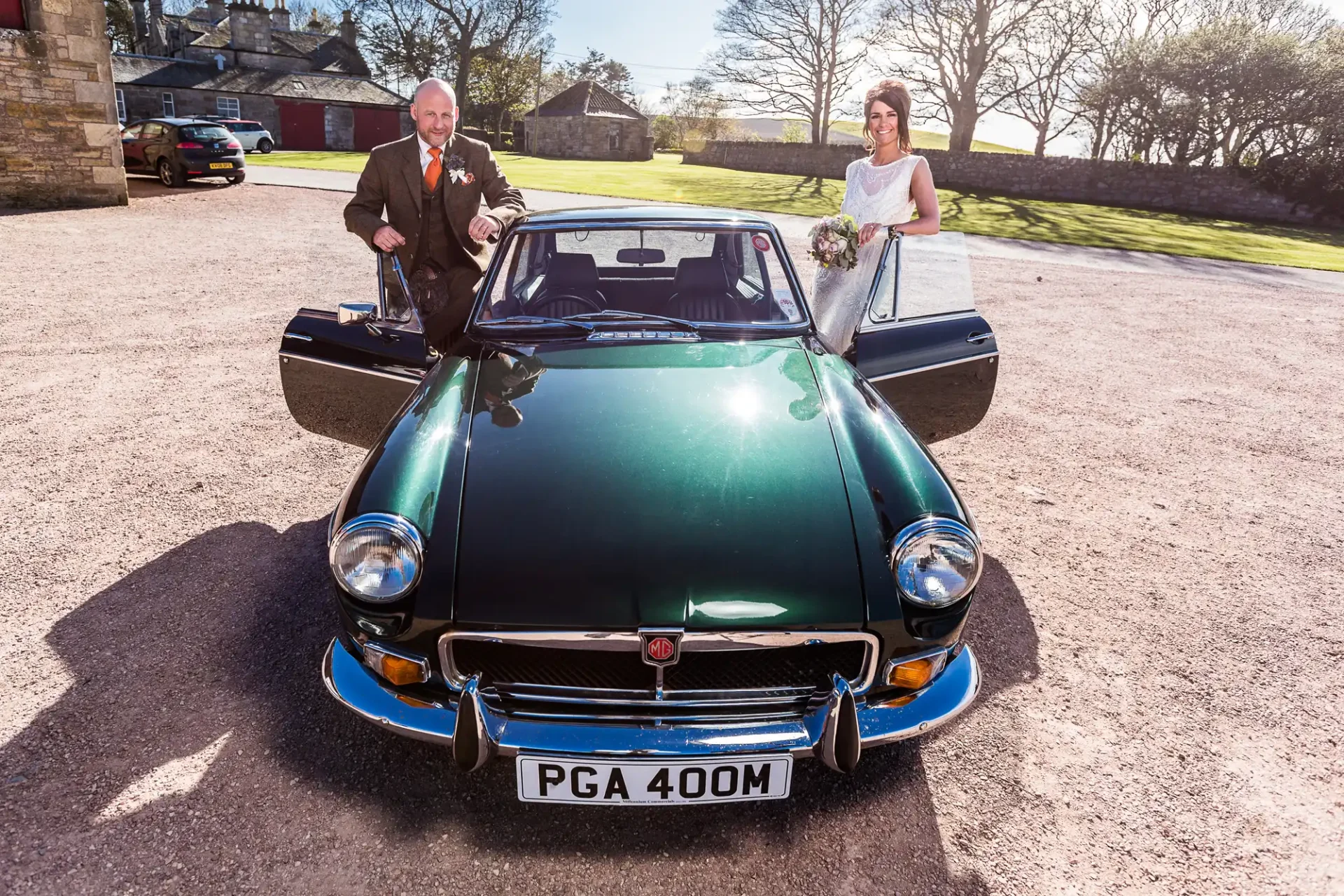A bride and groom standing by a green vintage car with open doors on a sunny day.