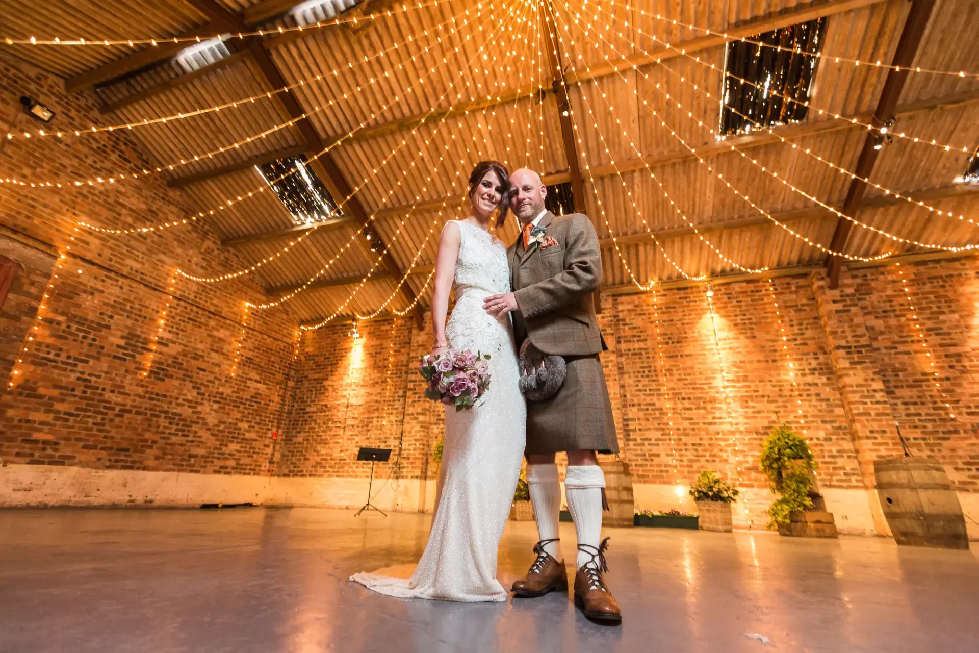 A bride and groom smiling while posing in a rustic hall adorned with fairy lights, the groom wearing traditional scottish kilt attire.