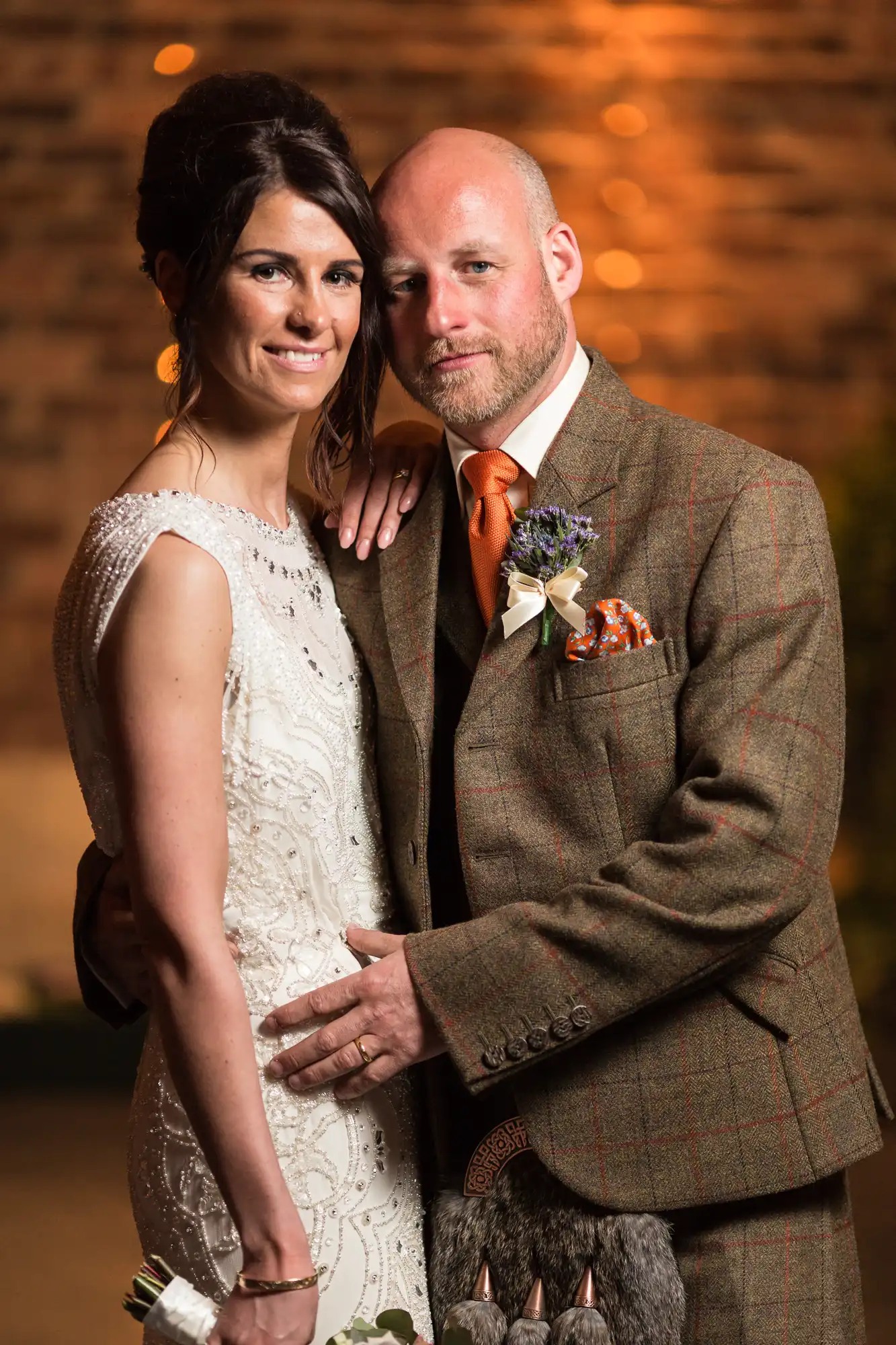 A bride and groom in formal wedding attire standing closely together with a warm, softly lit brick wall in the background.