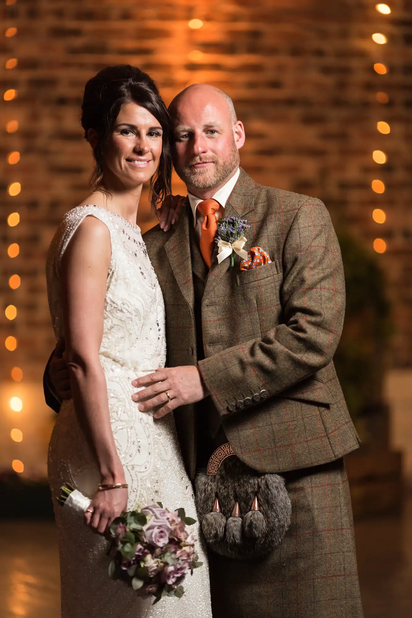 A bride in a lacy white dress and a groom in a tweed suit pose together, holding a bouquet, against a brick wall backdrop lit with warm lights.