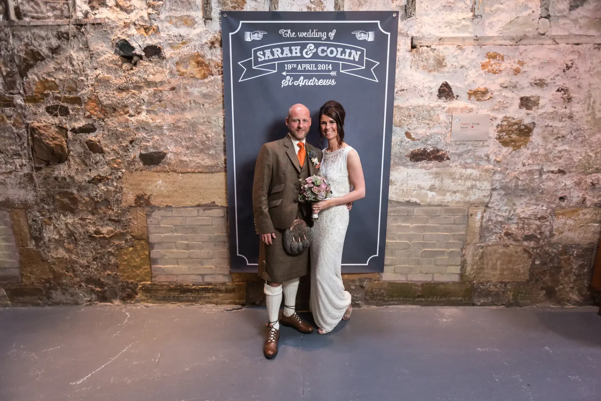 A bride and groom smiling in front of a decorative wedding sign with their names and wedding date, in a rustic indoor setting.