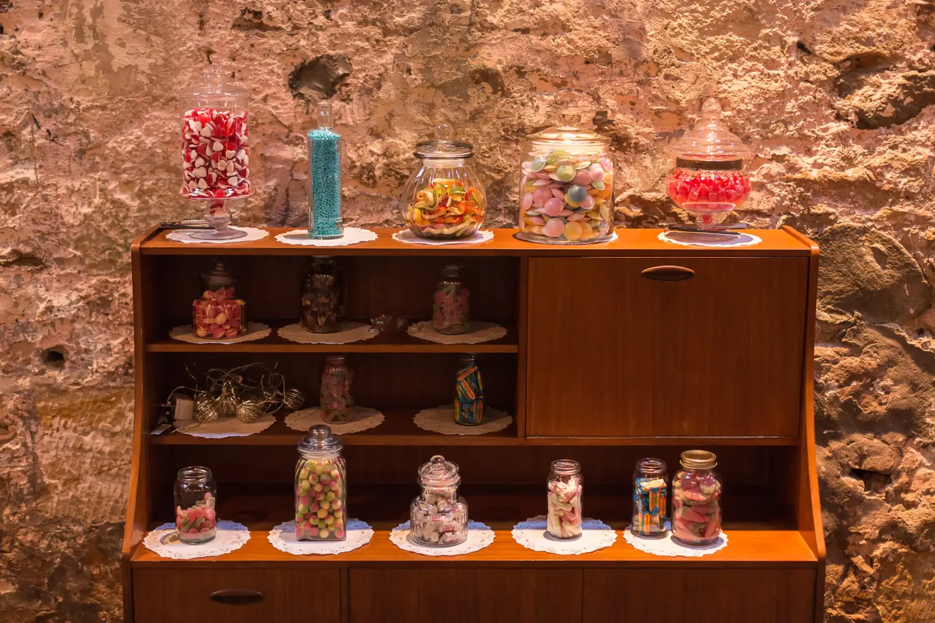 Various candies in glass jars displayed on a wooden shelf against a textured stone wall.