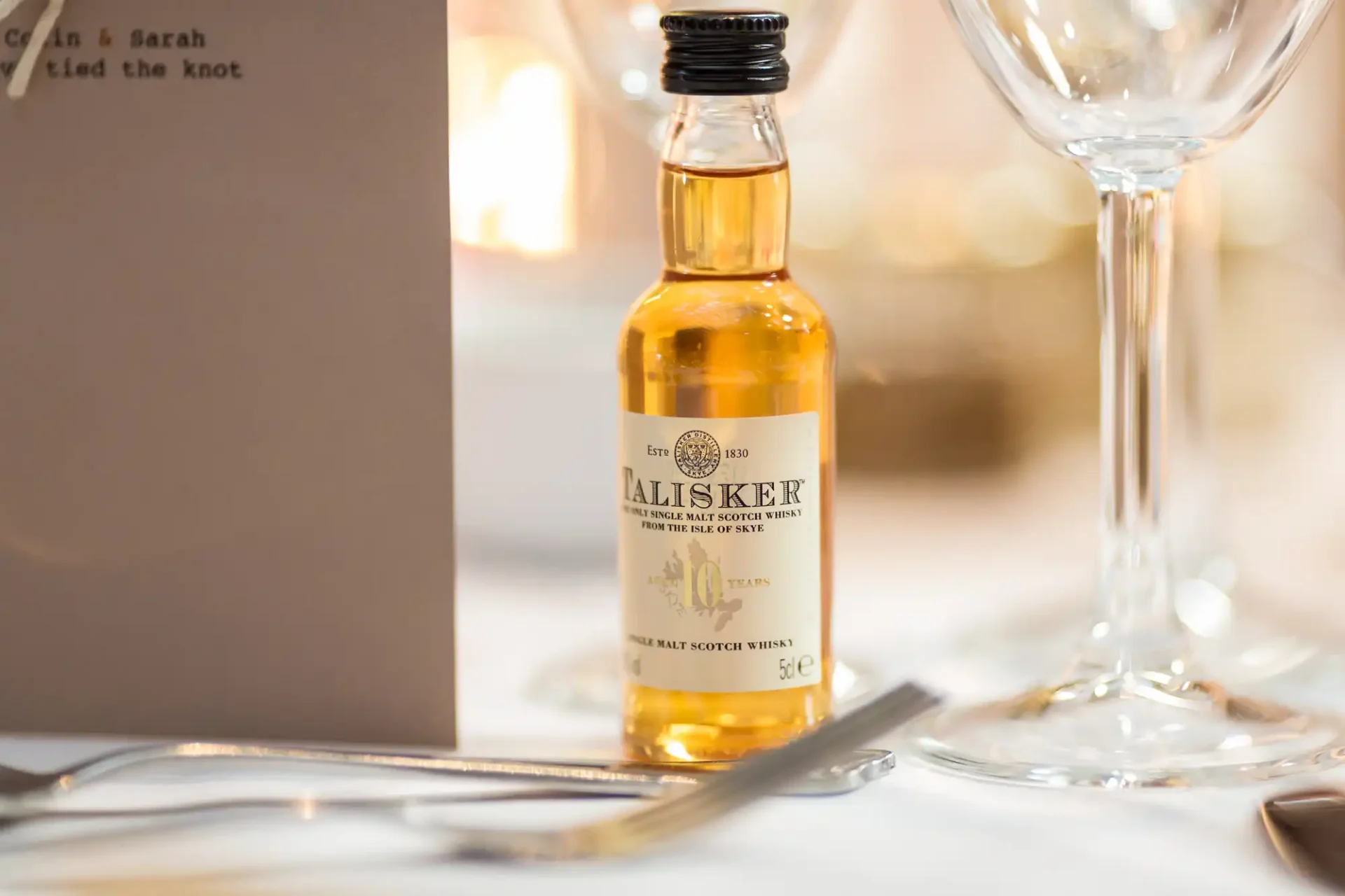 Small bottle of talisker scotch whisky on a wedding table, surrounded by elegant glassware and a menu card with names.