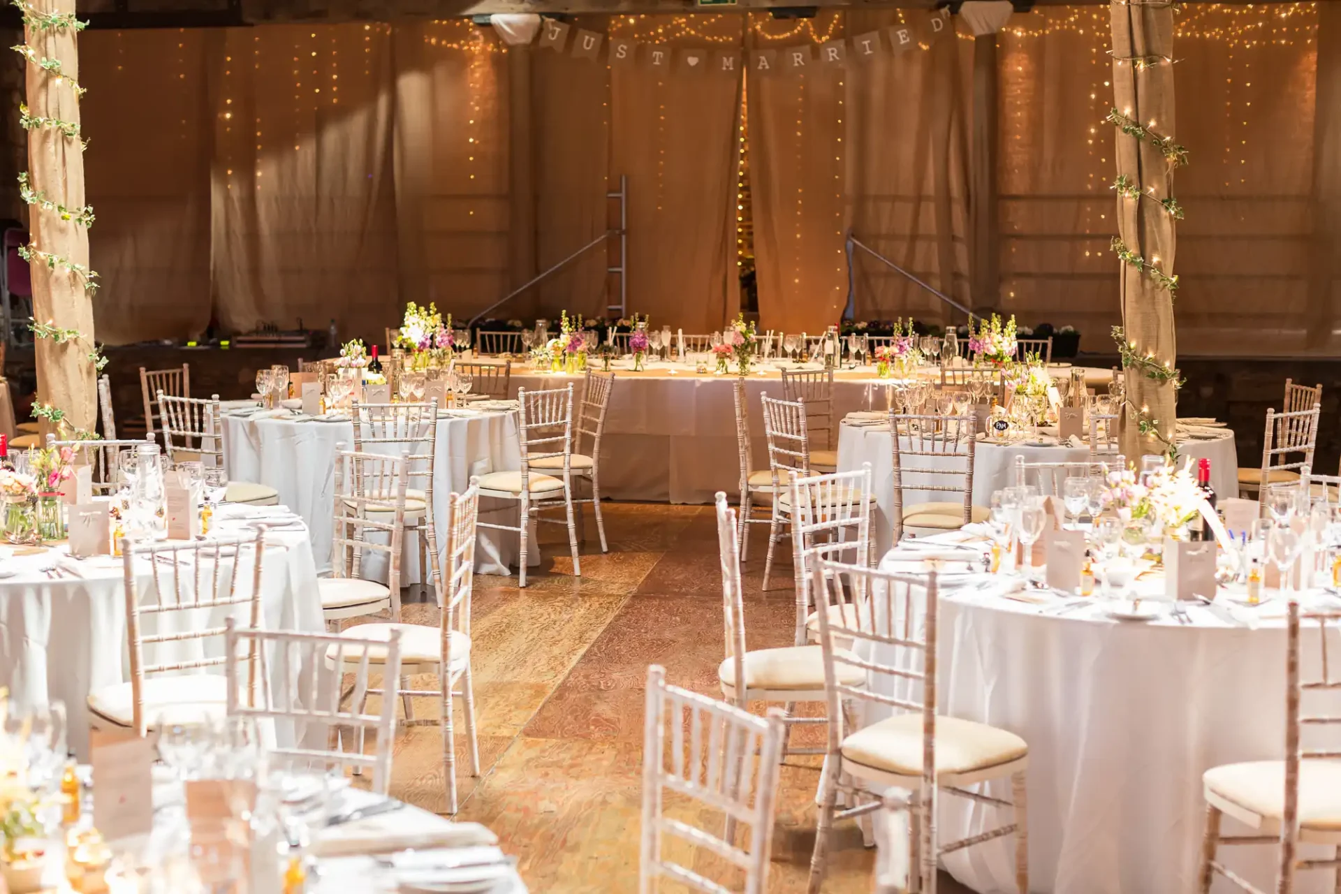 Elegant wedding reception hall decorated with white chairs, floral centerpieces, and string lights, with a "just married" sign in the background.