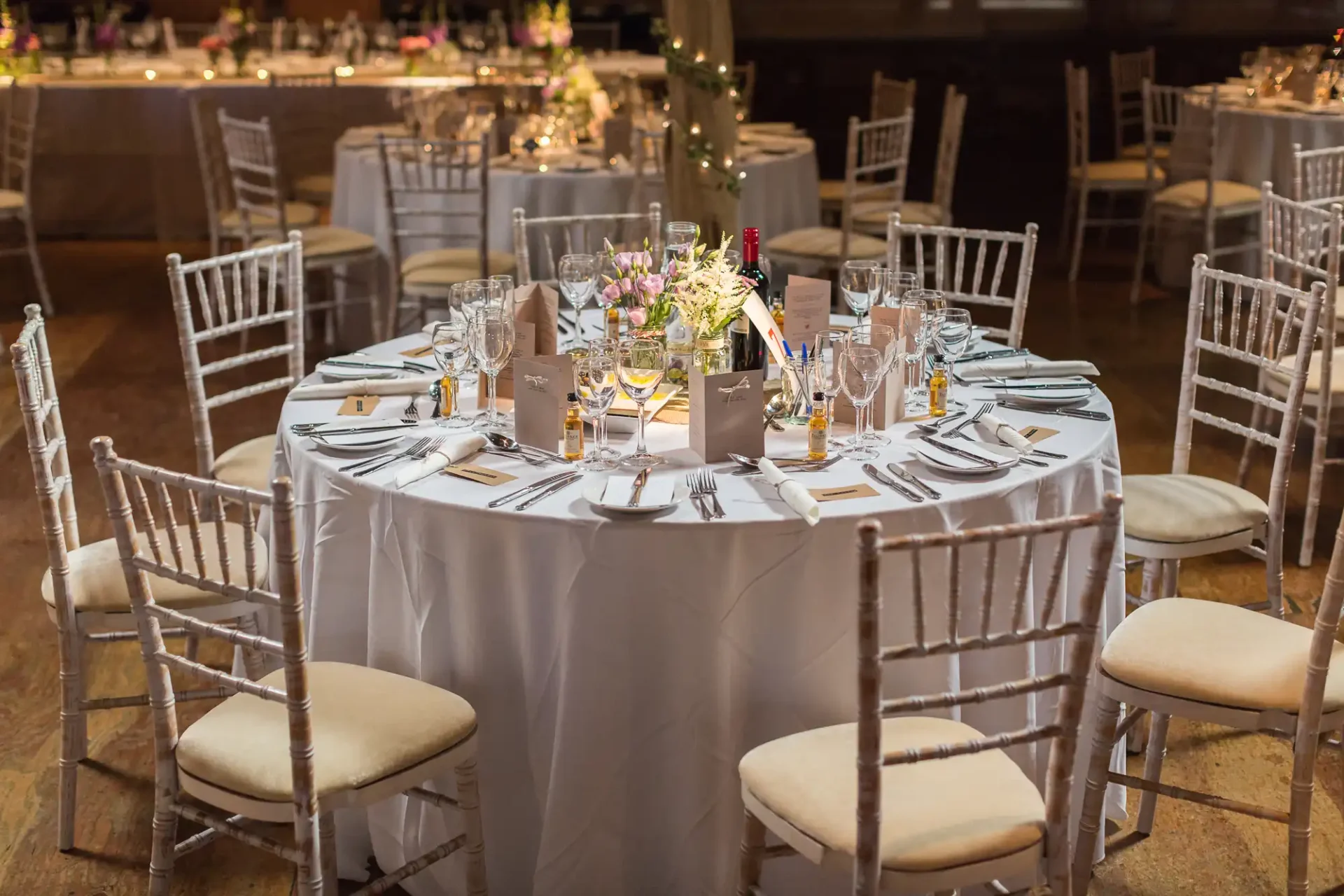 Elegant dining setup for a formal event with round tables, white linens, floral centerpieces, and chiavari chairs in a dimly lit venue.