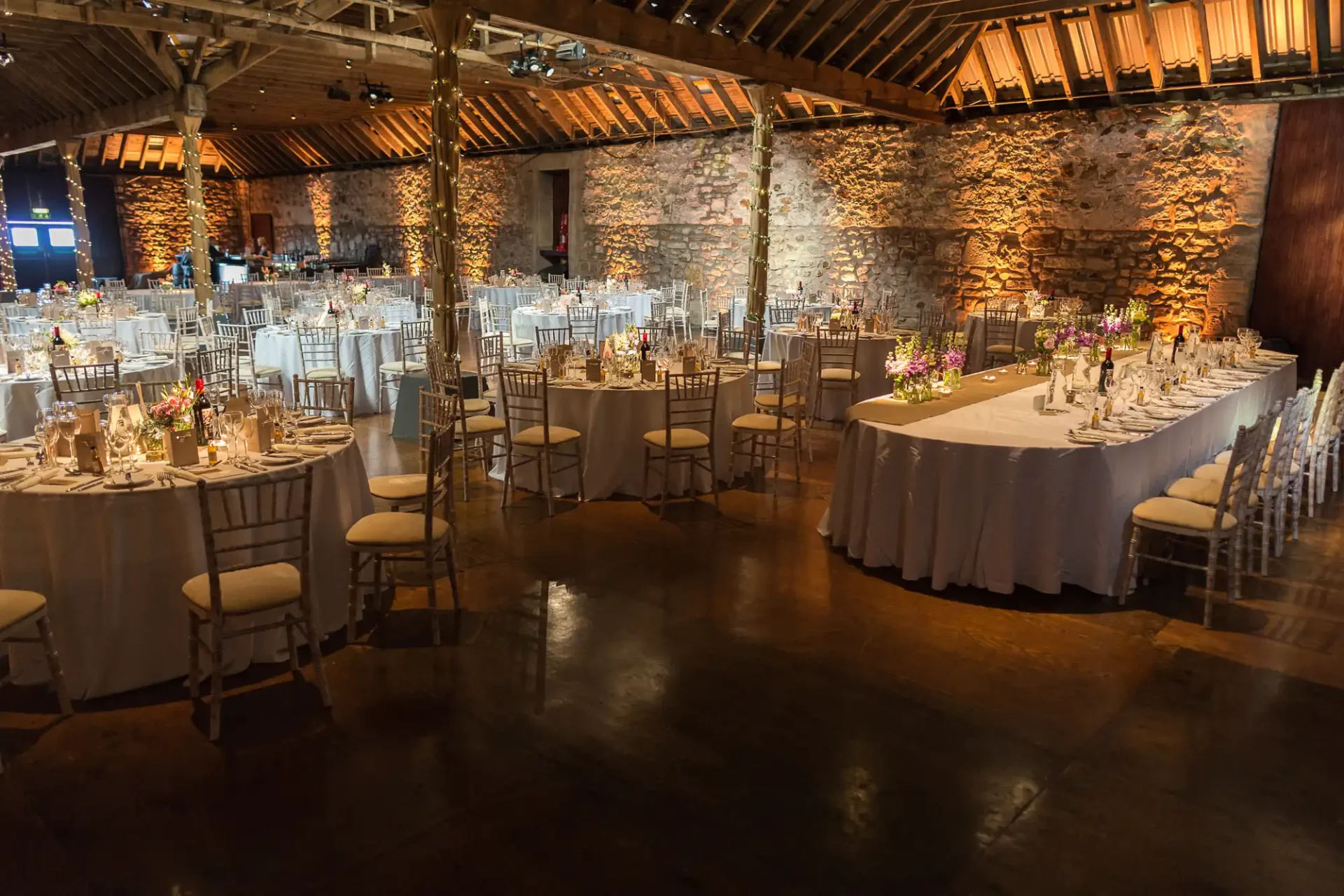Elegant dining setup in a rustic venue with stone walls and wooden ceiling, featuring round tables with white linens and chairs.
