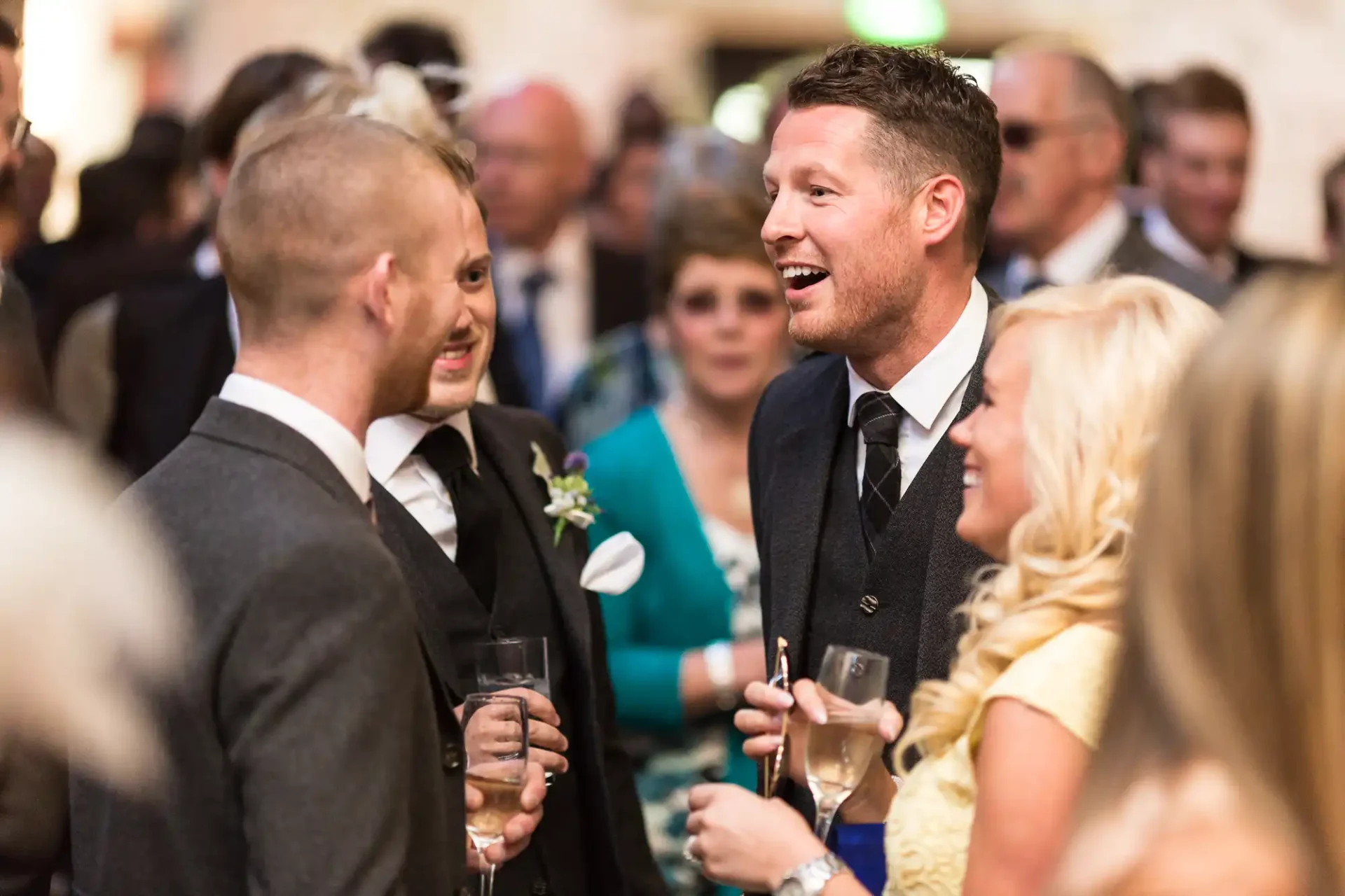 Two men in suits smiling and talking at a wedding reception, surrounded by guests with drinks.