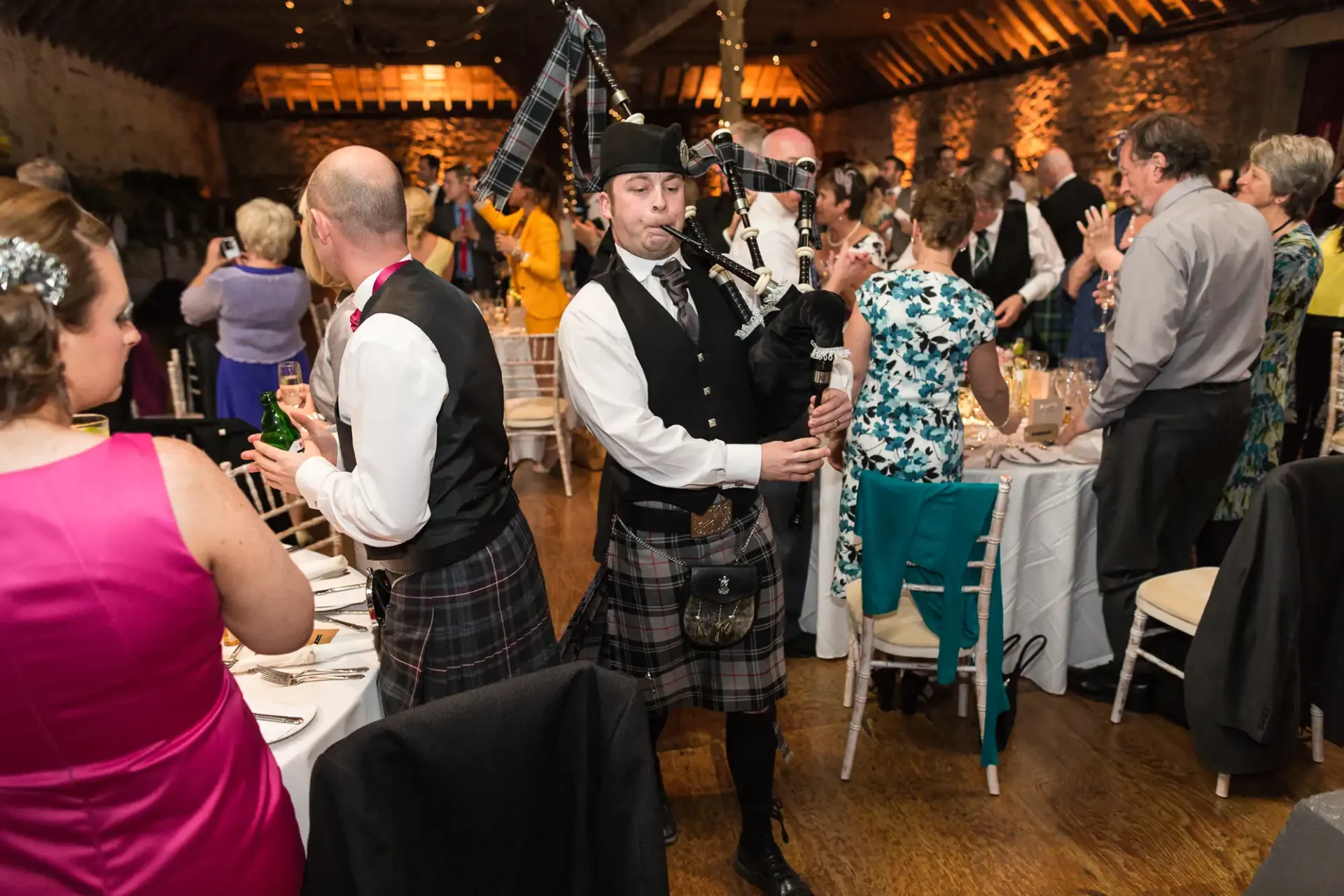 A bagpiper in traditional scottish attire performing at a lively indoor wedding reception with guests mingling around tables.