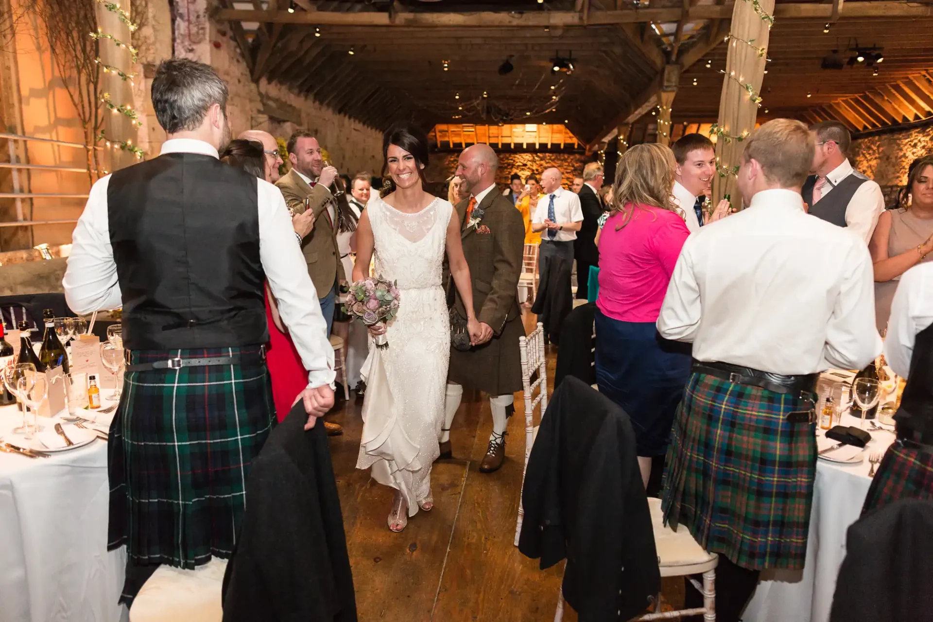 A bride and groom smiling as they walk through a crowd of guests at a rustic wedding reception, with many men wearing kilts.