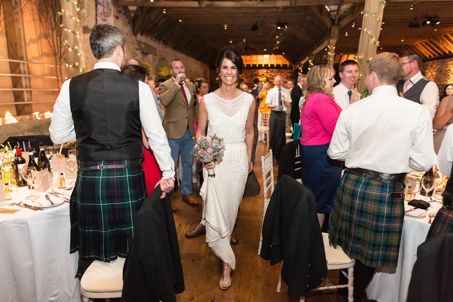 Bride walking through wedding reception hall, guests clapping, two men in kilts facing her. warm lighting and rustic decor.
