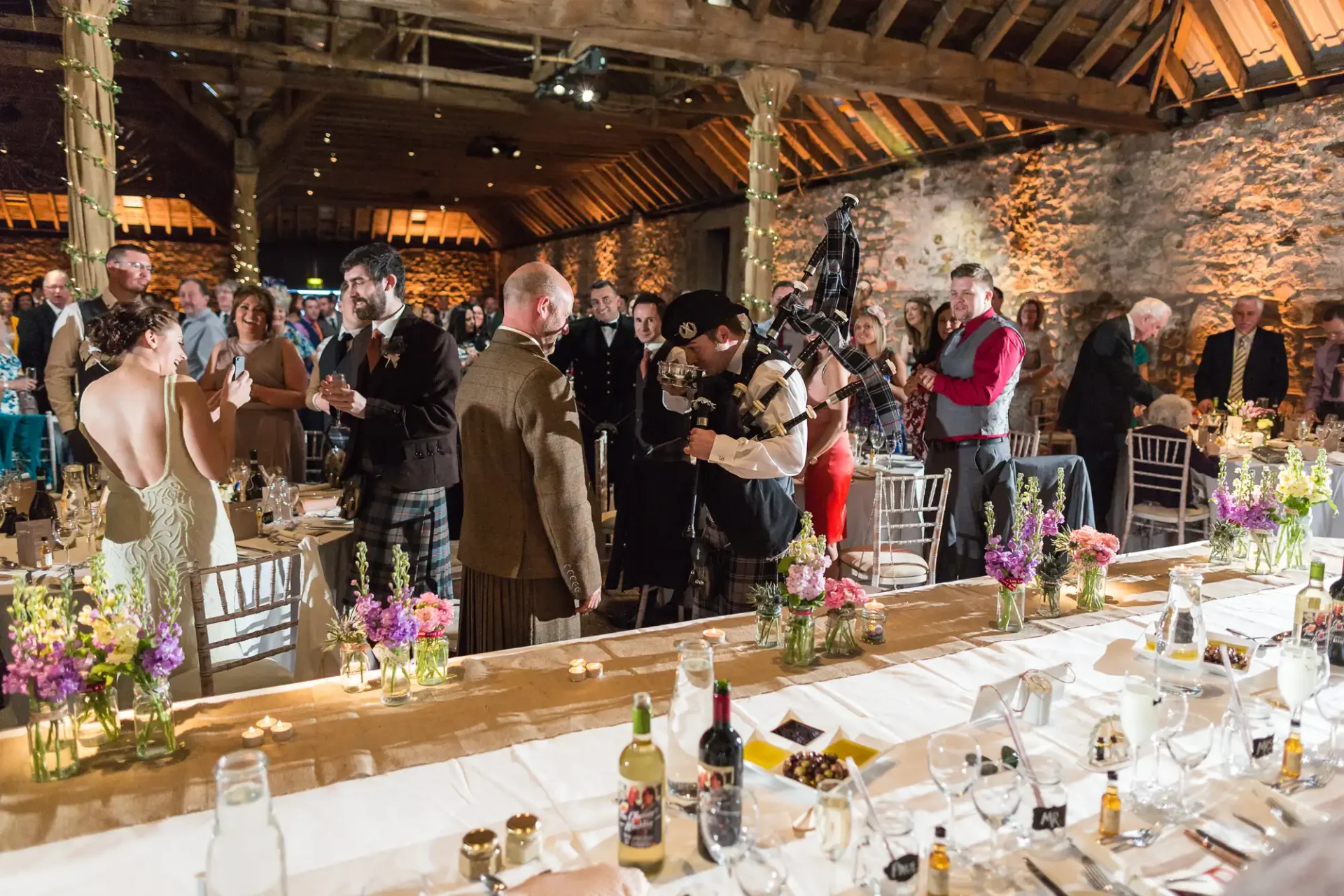 A lively wedding reception inside a rustic hall, with guests clapping for a bagpiper walking past decorated tables.
