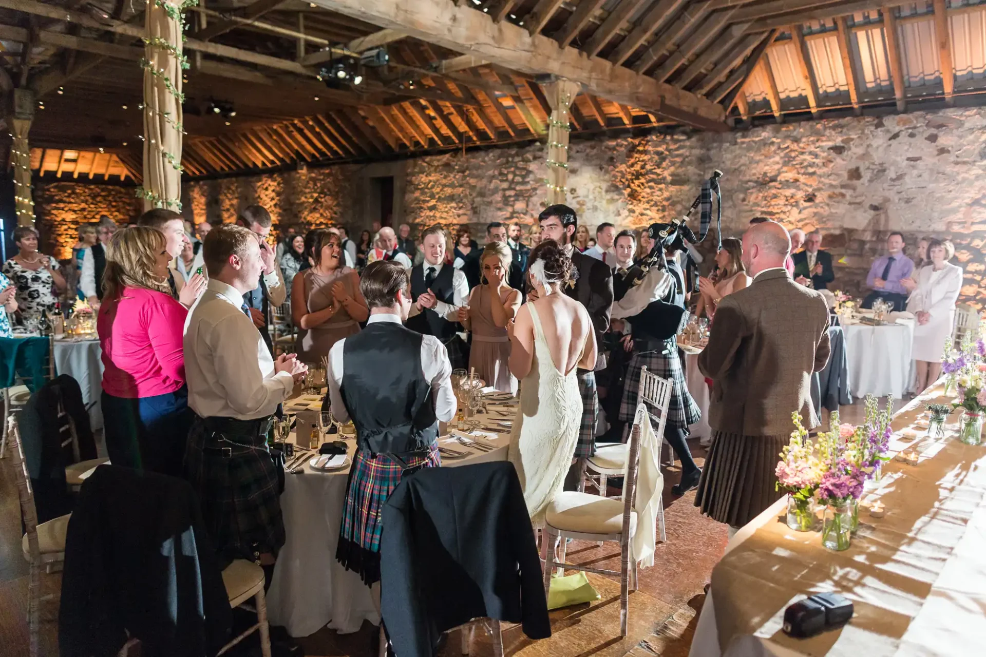 A wedding reception in a rustic stone barn, with guests standing and applauding the bride and groom at the head table.