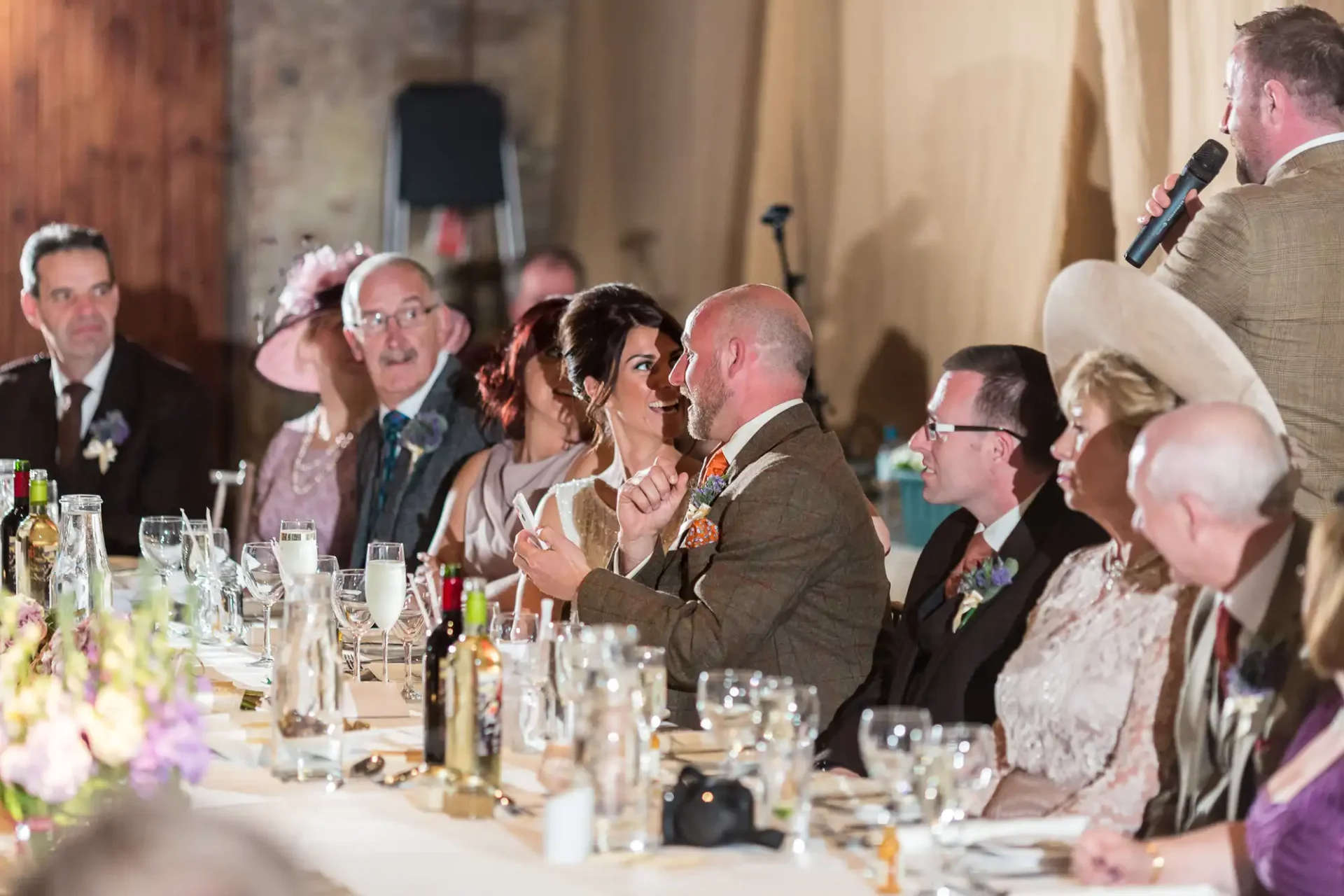 A wedding reception scene with a bride and groom seated at a long table, conversing and smiling, while a man standing delivers a speech with a microphone.