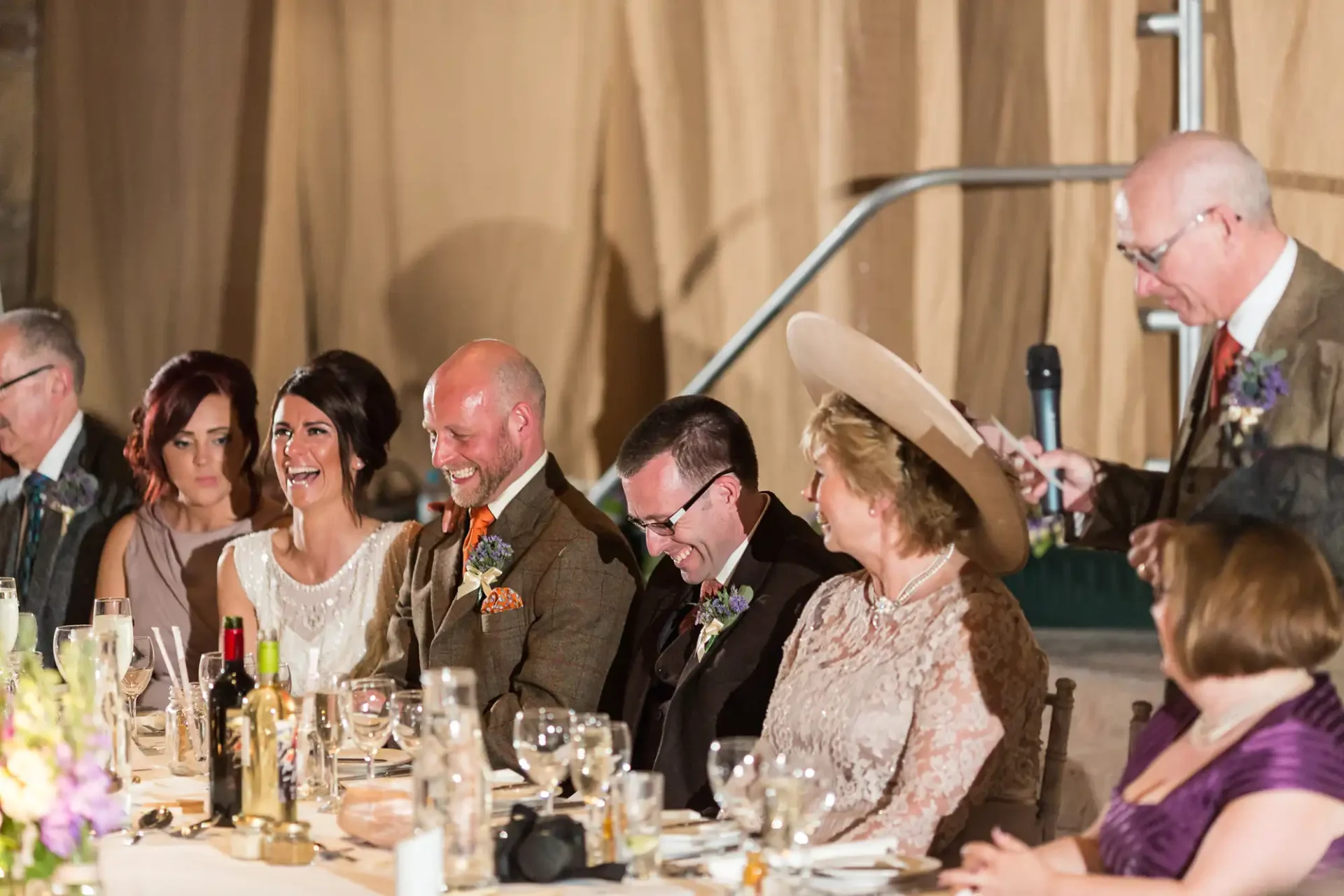 A joyful wedding reception scene with guests laughing around a dinner table, elegantly dressed, including a bride and groom.