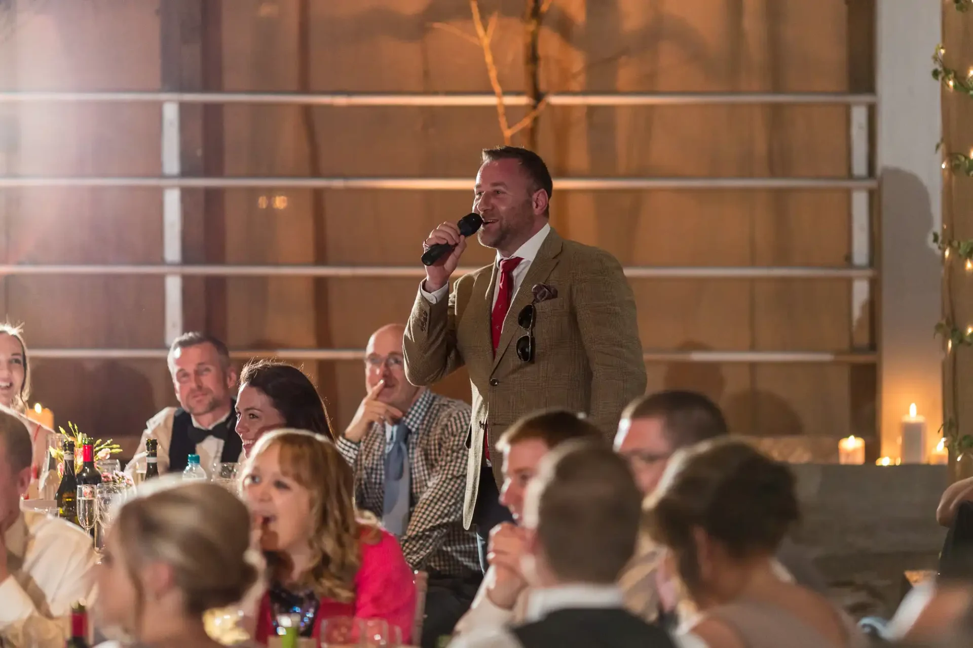 A man in a tweed jacket and red boutonniere speaks into a microphone at a festive event, with people seated at tables in soft-lit background.