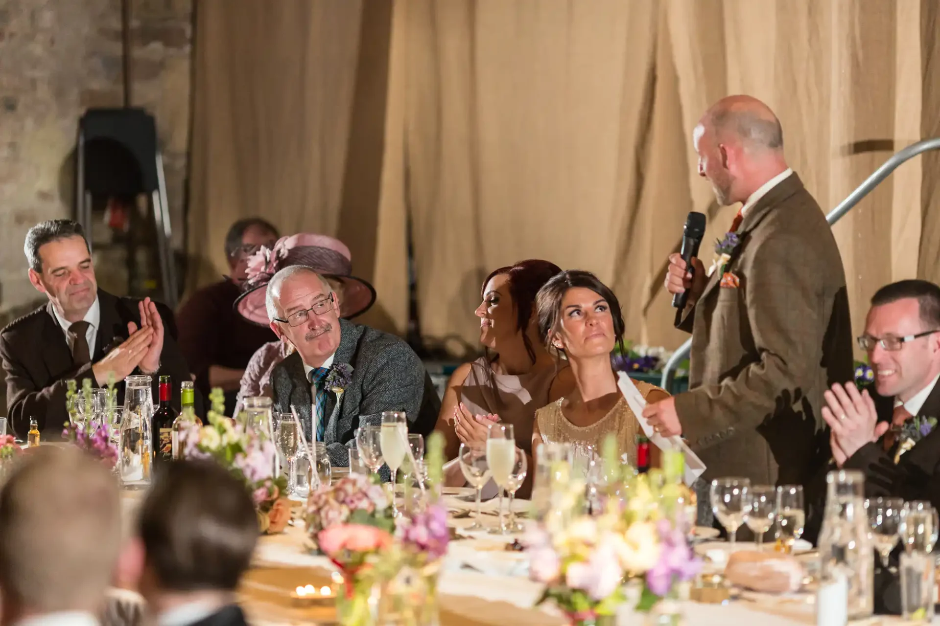 A man gives a speech at a wedding reception, holding a microphone, with guests seated at a table listening attentively, some clapping.