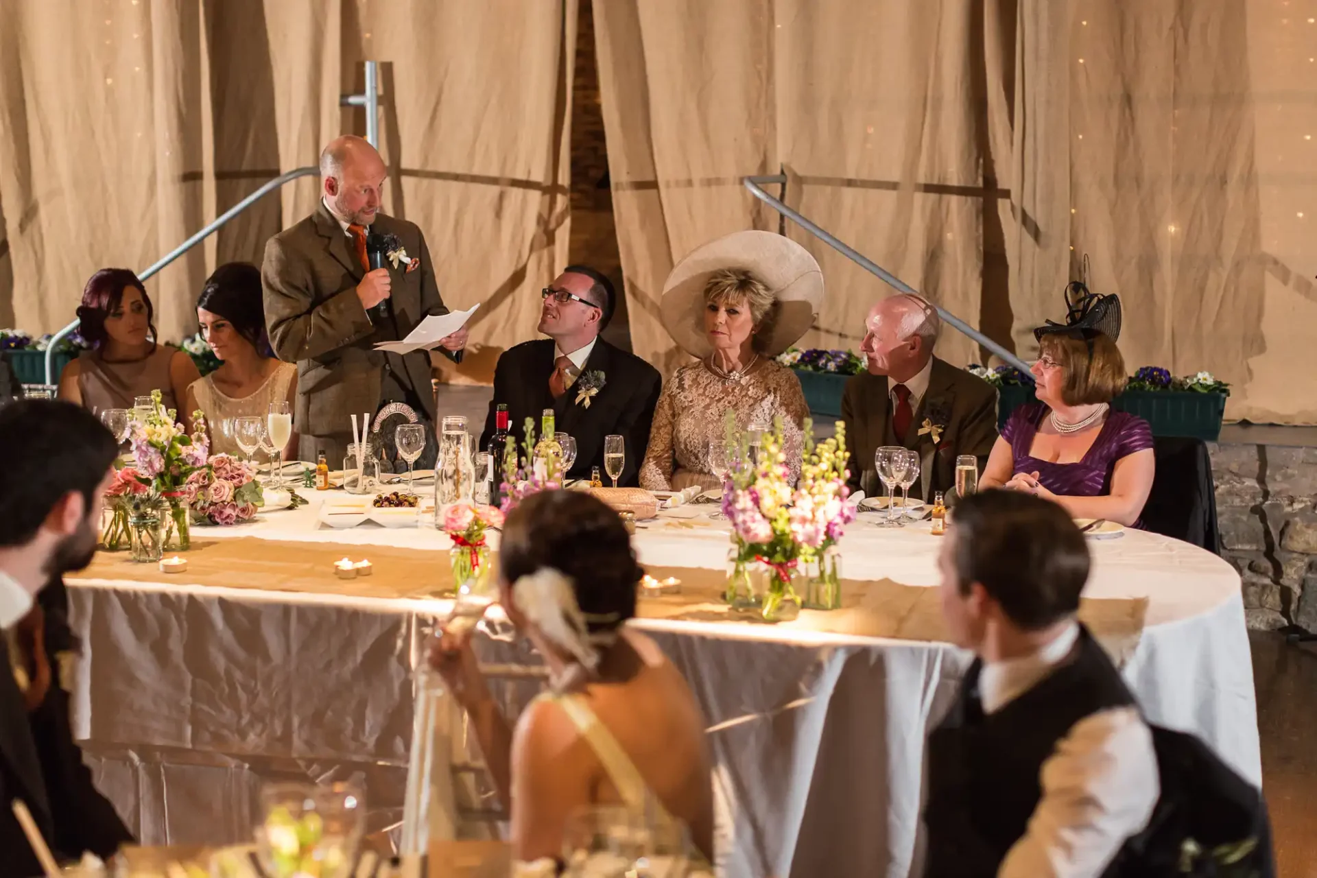 A man gives a speech at a wedding reception, holding a microphone, with guests seated around decorated tables listening attentively.