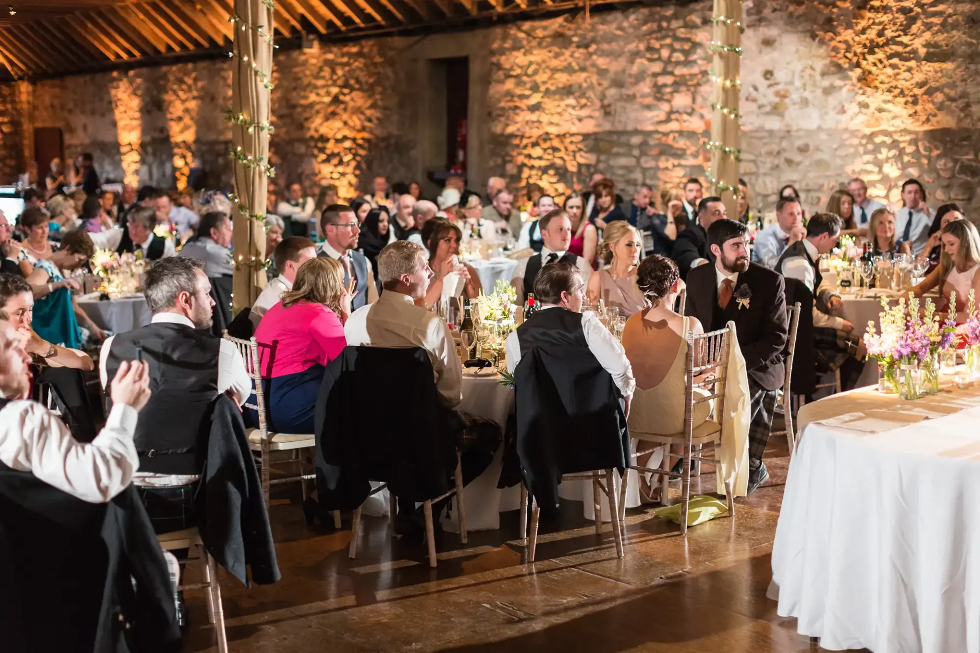 Guests seated at long tables during a wedding reception in a rustic stone hall, decorated with flowers and candles.