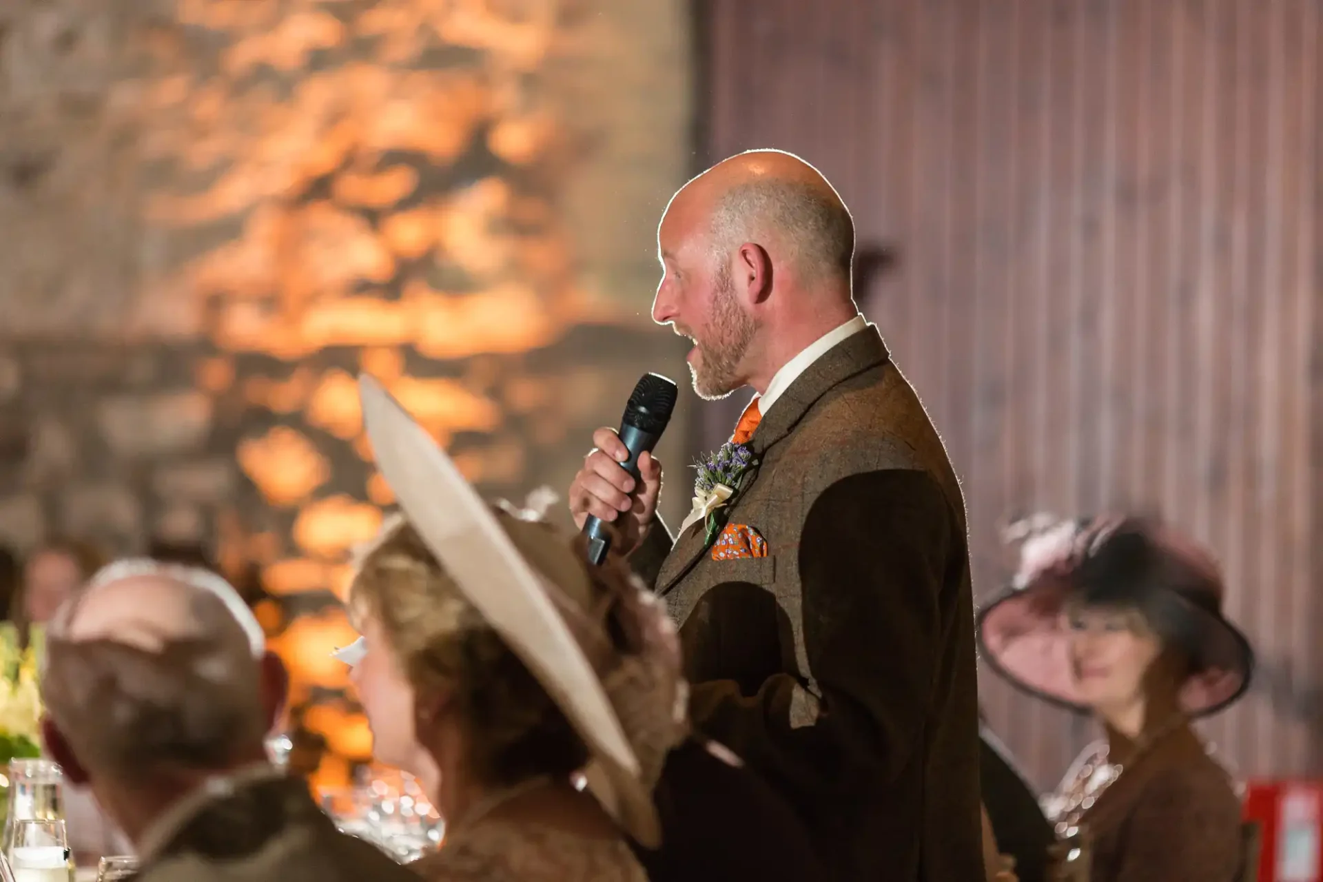 Bald man in a tweed suit giving a speech at a wedding, holding a microphone and a paper, with guests in fancy hats blurred in the background.