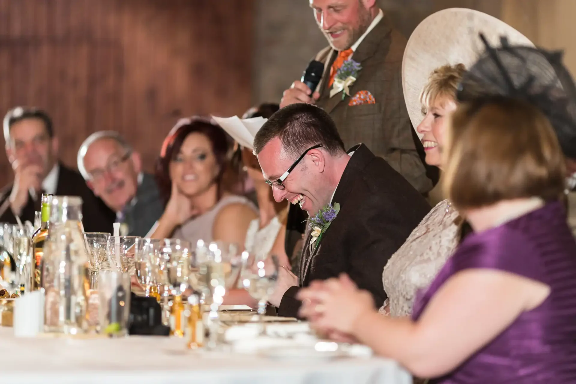 A man in a kippah laughs heartily at a wedding table while others, including a man with a microphone, enjoy the moment.