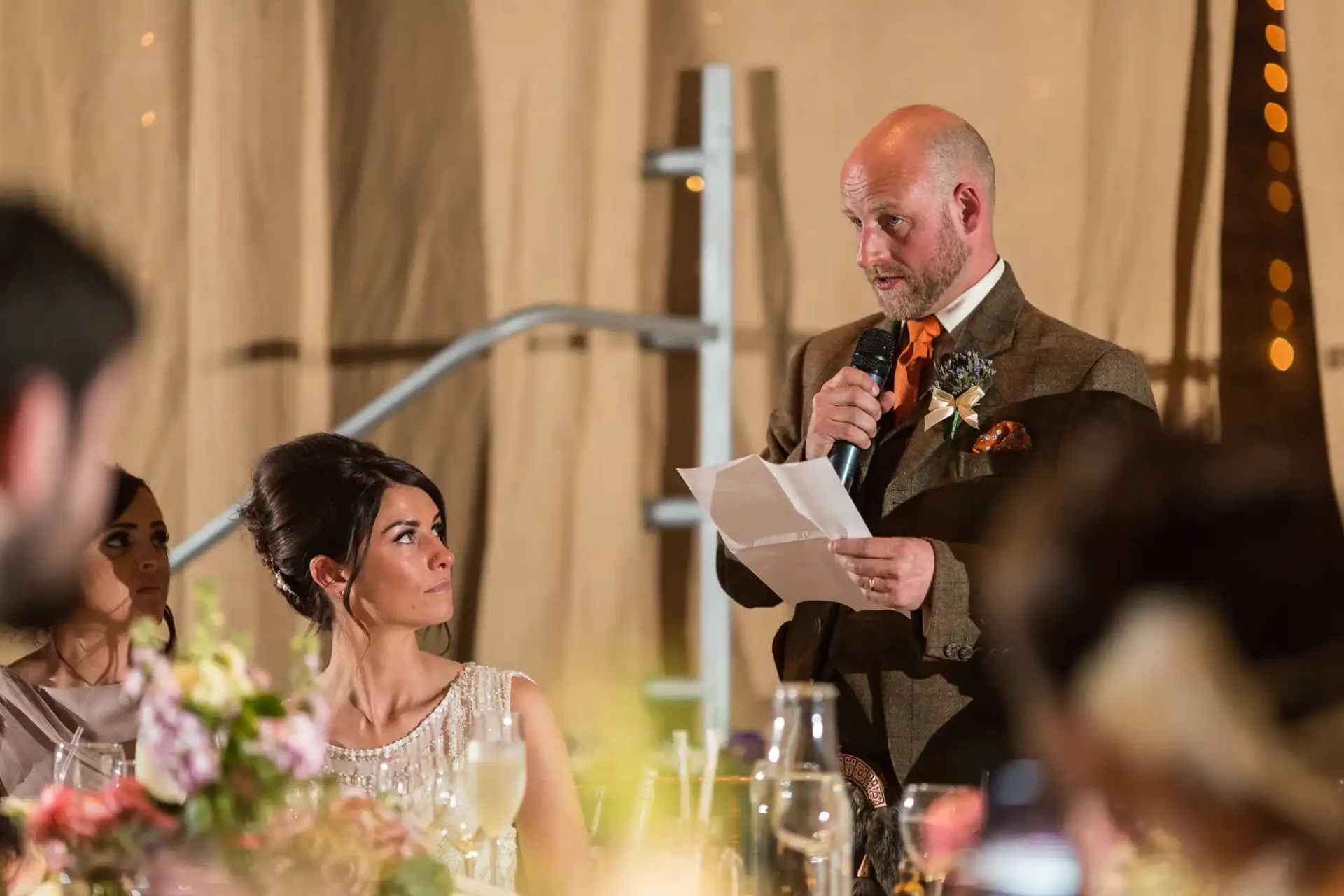 Bald man in a tweed suit delivers a speech with a microphone at a wedding reception, reading from a paper, with a focused woman in the foreground.