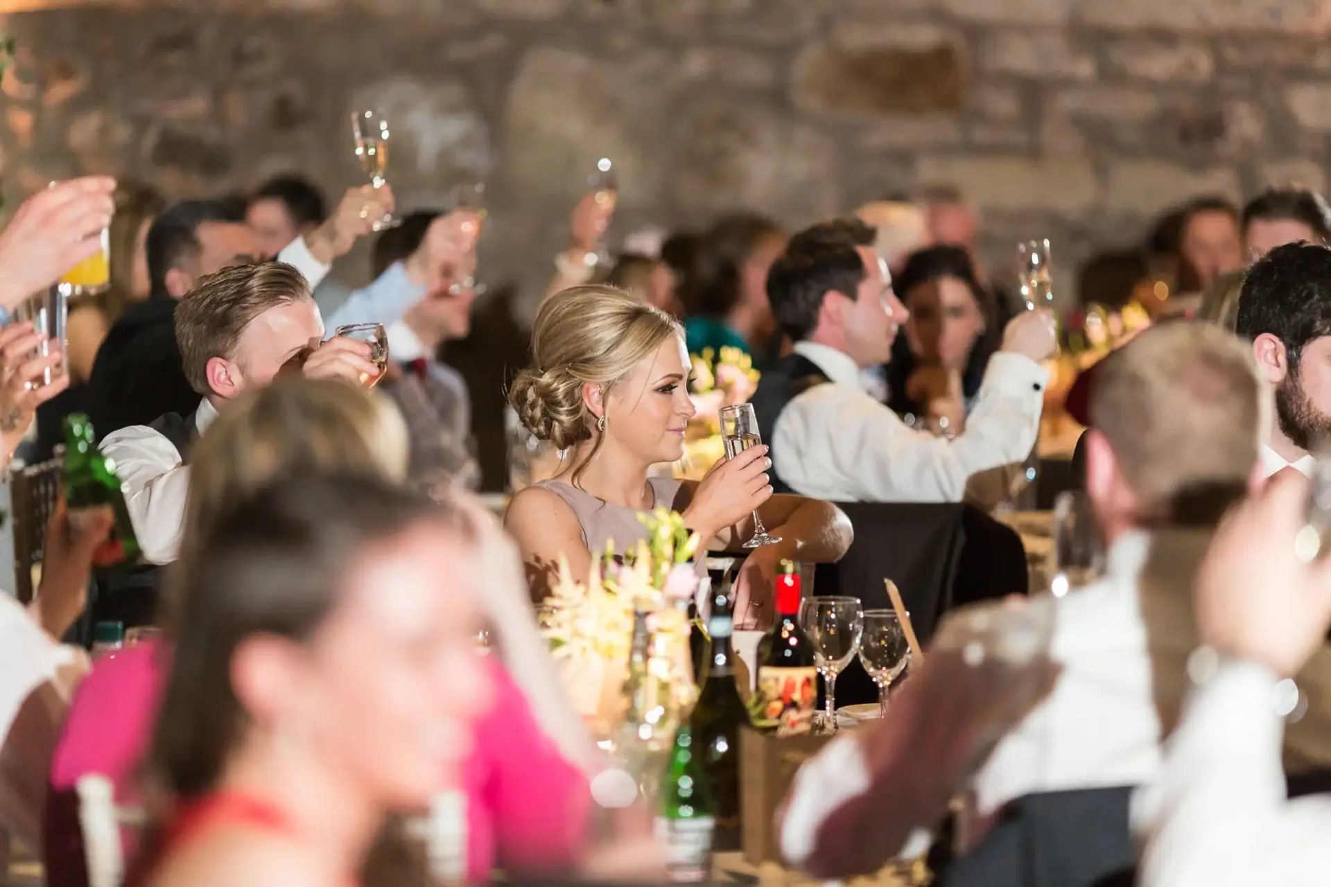 Guests raising glasses for a toast at a candlelit wedding reception inside a stone-walled venue.