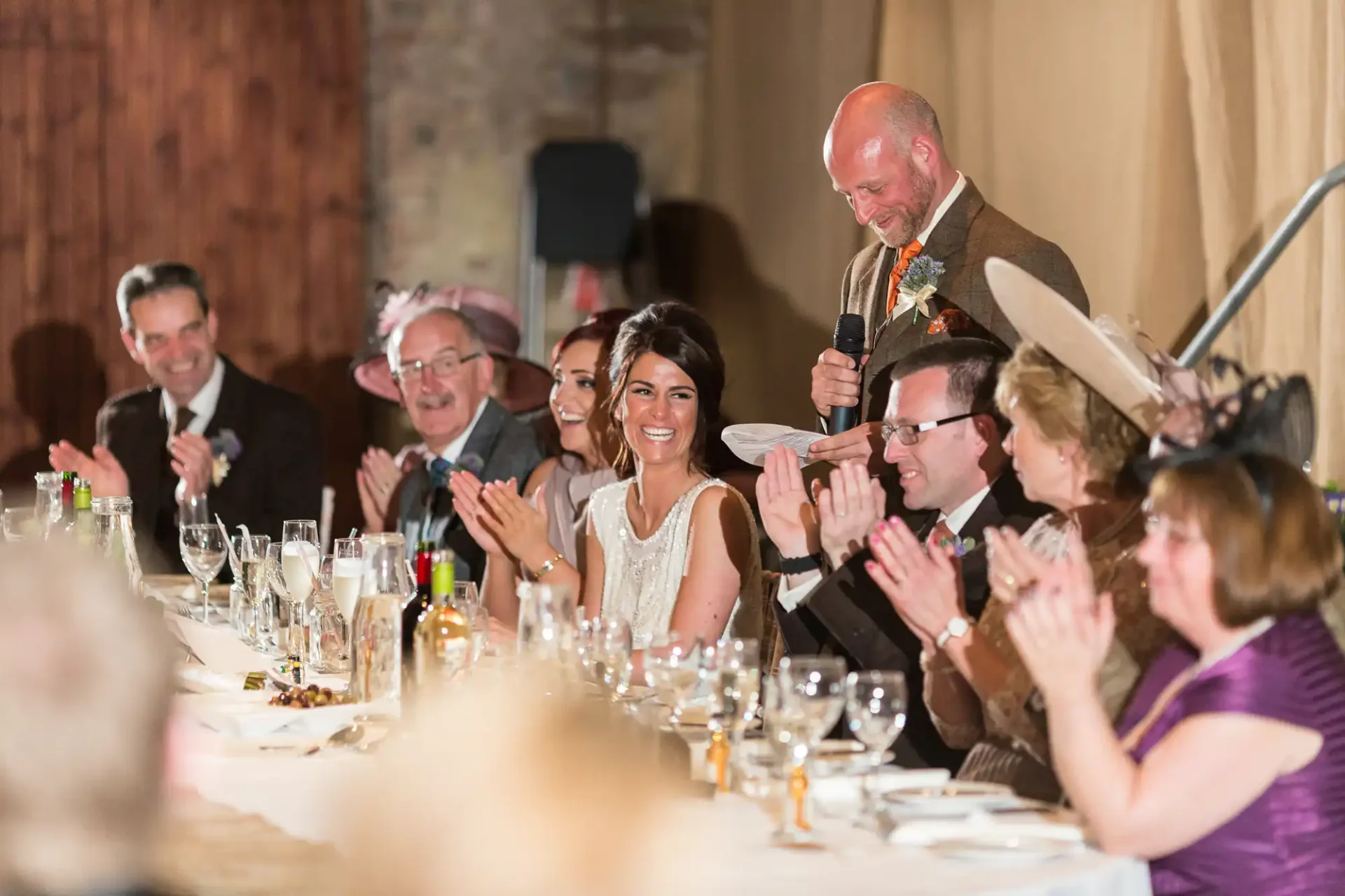 A joyful wedding reception scene with guests clapping as a bald man shares a laugh, while a woman beside him smiles widely at a festively decorated table.