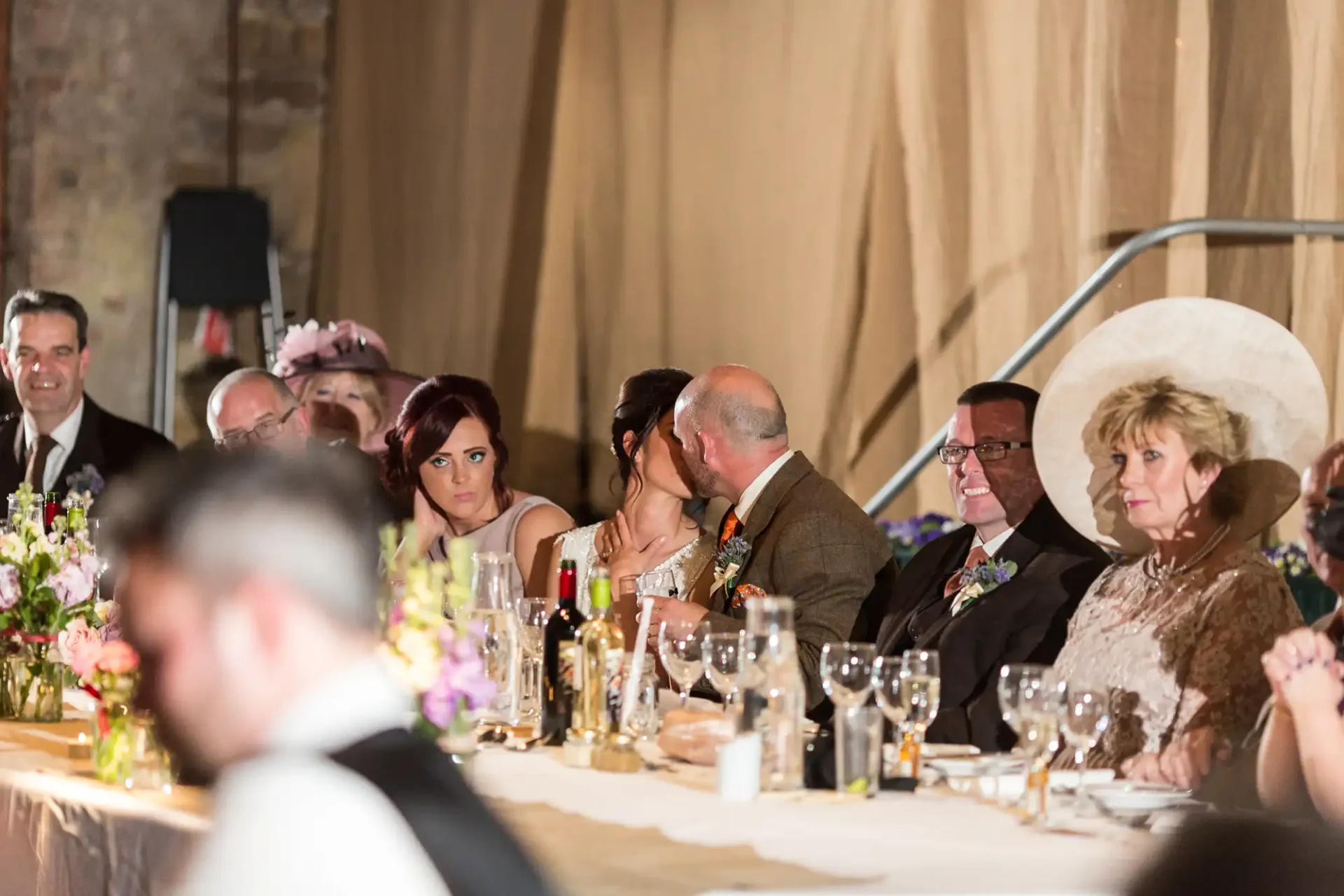 A couple kissing at a wedding reception table surrounded by guests, with floral centerpieces and candles, in a warmly lit venue.