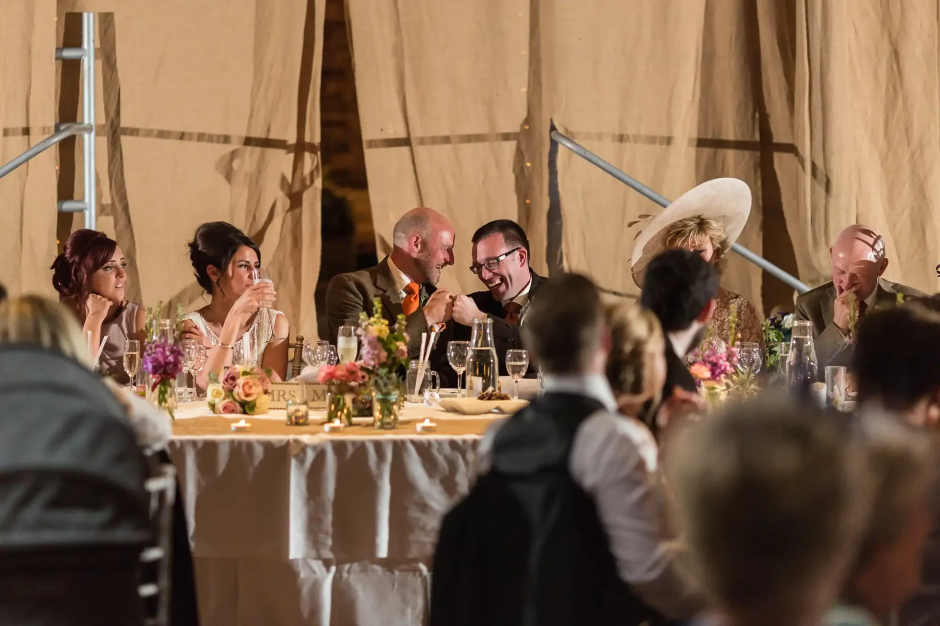 Wedding guests laughing and interacting at a banquet table decorated with flowers inside a tent.