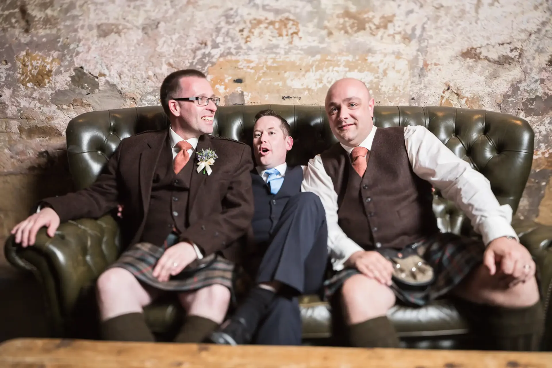 Three men in kilts and vests sitting on a leather sofa laughing, one with a visible wedding boutonniere.
