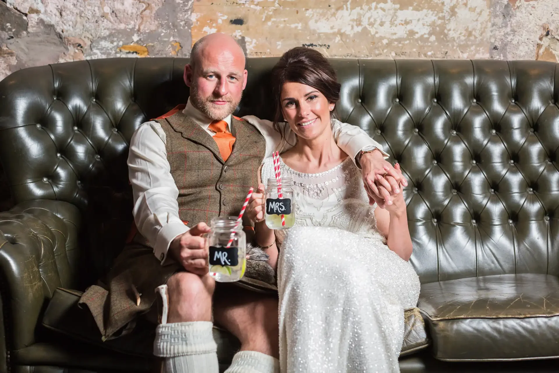 A couple in wedding attire sits on a leather sofa, smiling and holding mugs labelled "mr." and "mrs." in a rustic setting.
