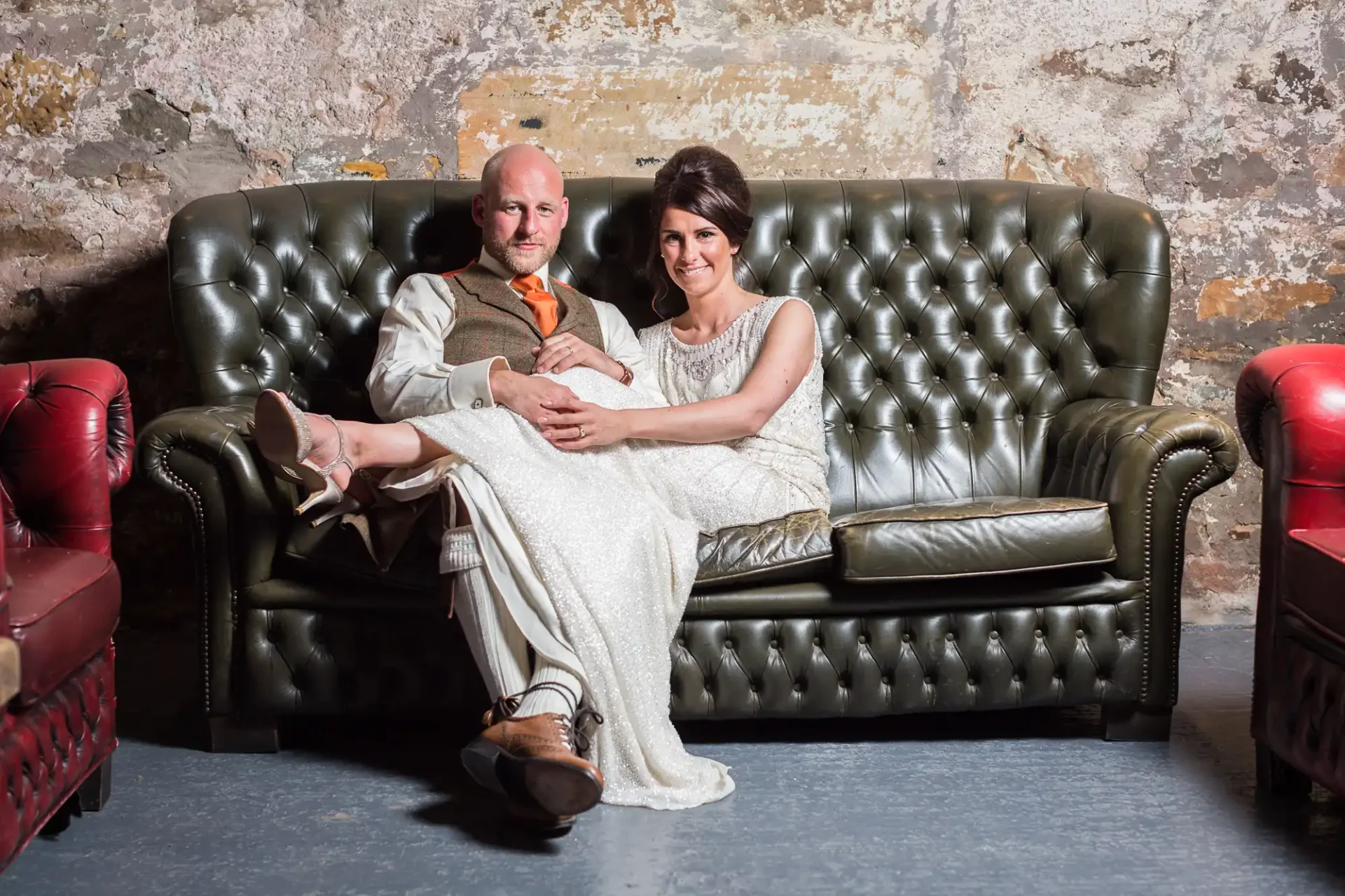 A bald man in a suit and a woman in a long dress sitting closely on a vintage leather sofa, looking at the camera, with textured walls in the background.